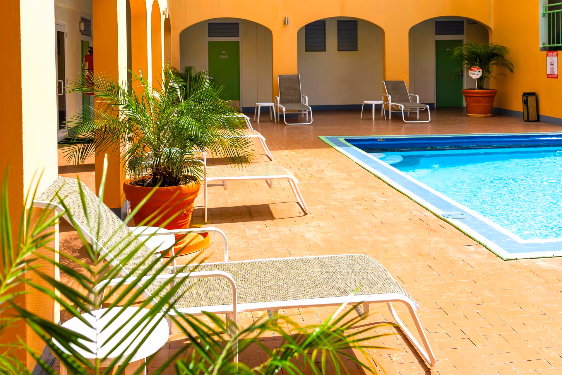 Outdoor pool area with blue water, surrounded by yellow walls, potted plants, and multiple white lounge chairs with side tables.