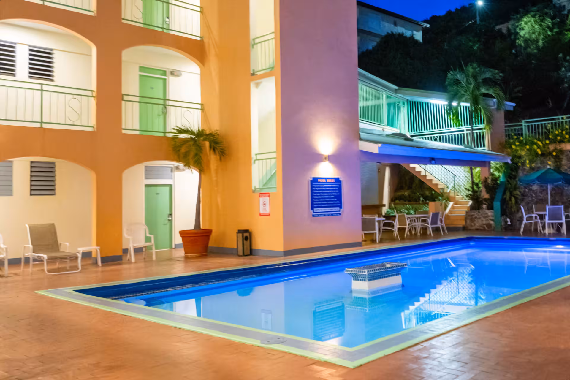 Well-lit outdoor swimming pool at night surrounded by lounge chairs, palm trees, and a multi-story building with green doors and railings.
