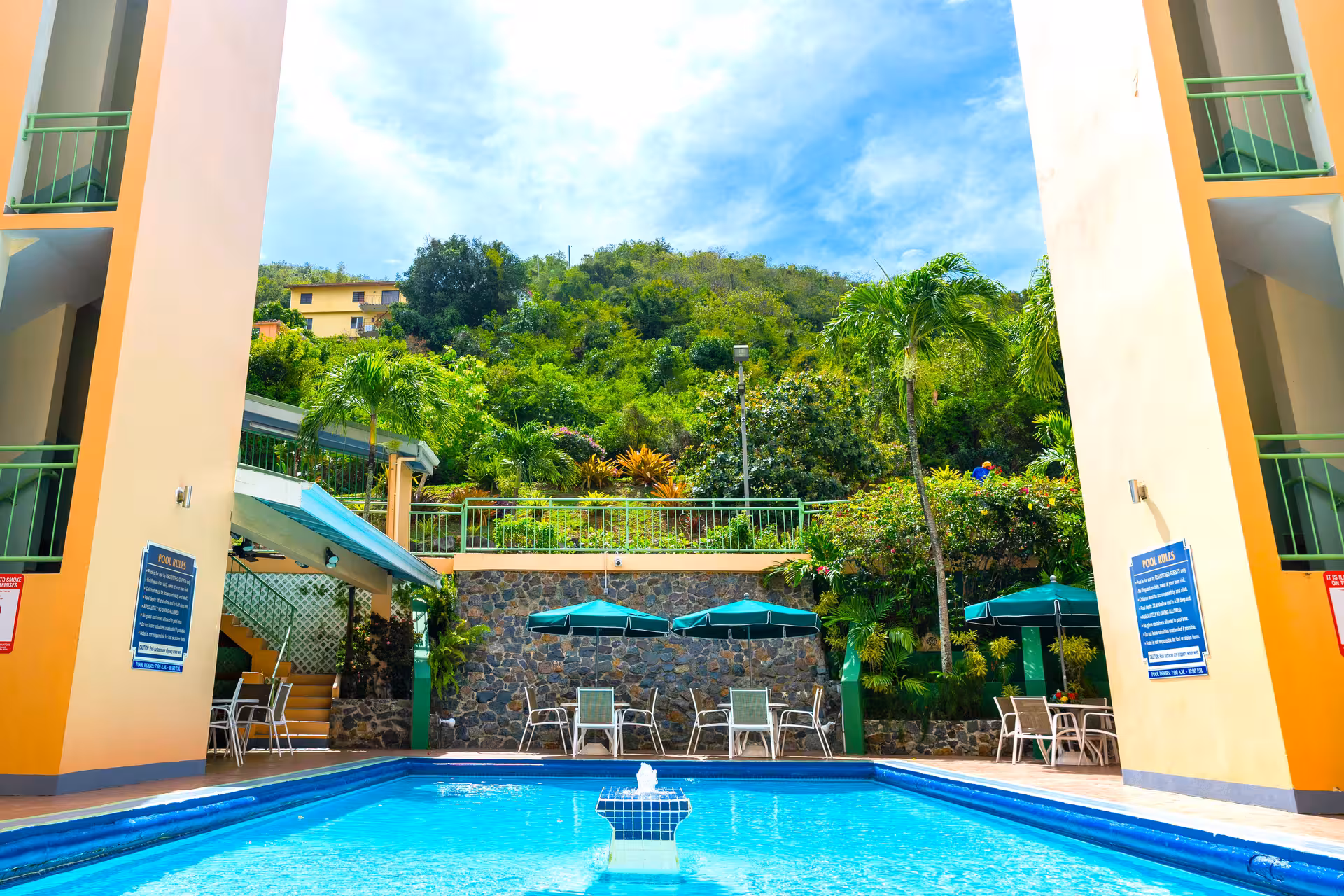 Bright outdoor swimming pool with blue water, surrounded by yellow columns, green umbrellas, chairs, palm trees, and lush greenery.