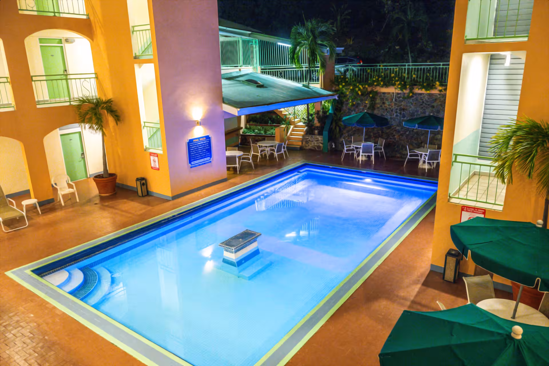 Well-lit rectangular outdoor swimming pool surrounded by yellow building walls, green umbrellas, white chairs, and tables at night.