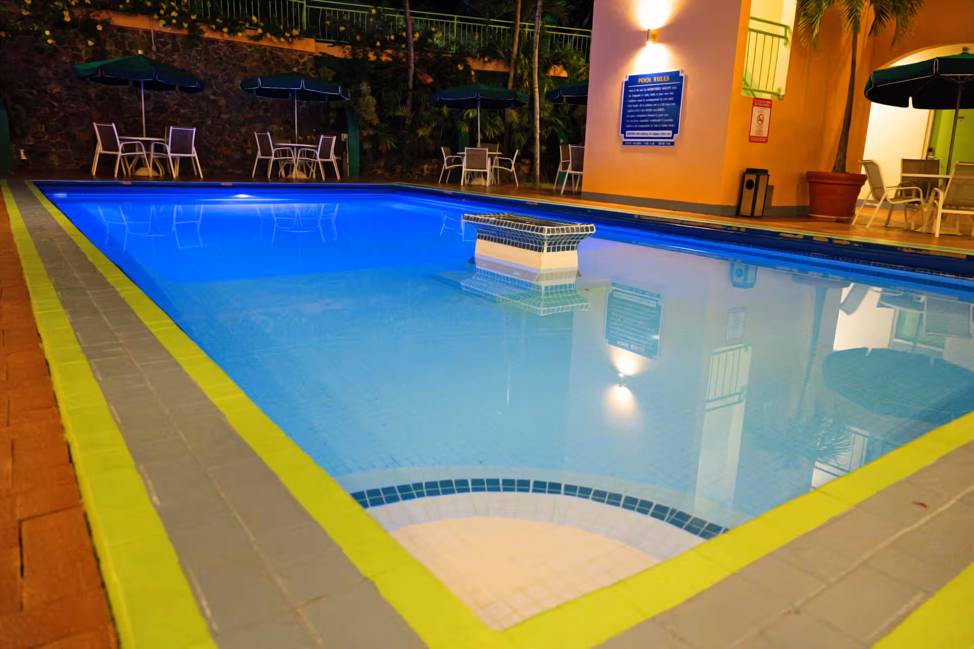 Clean outdoor swimming pool with bright blue water at night, surrounded by tables with umbrellas and chairs.