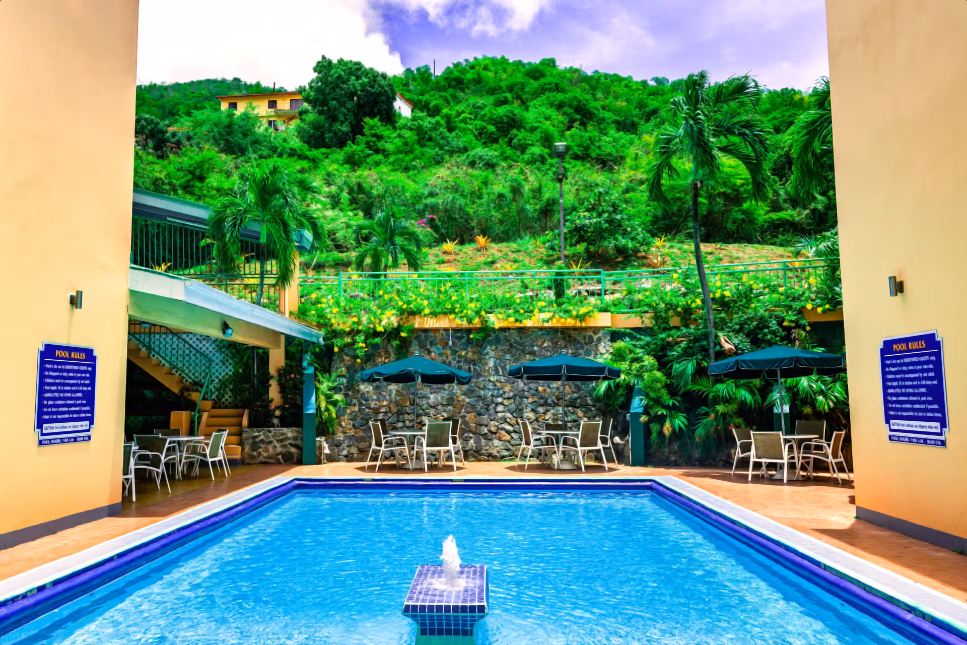 Clear blue swimming pool with central water fountain, surrounded by patio tables with umbrellas and lush green hillside in the background.
