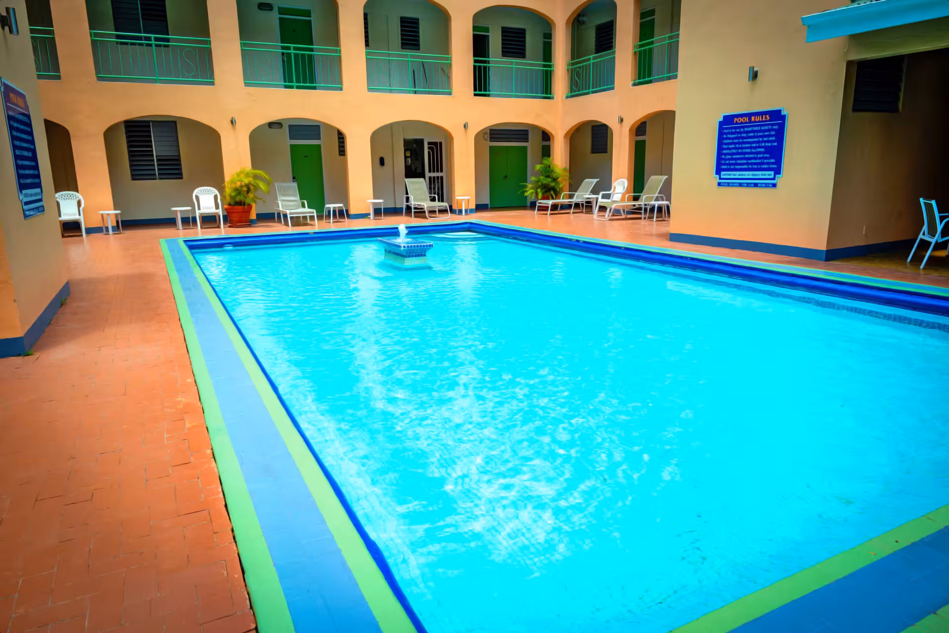 Indoor rectangular swimming pool with blue water and a small fountain in the center, surrounded by lounge chairs and green doors in a peach-colored building.