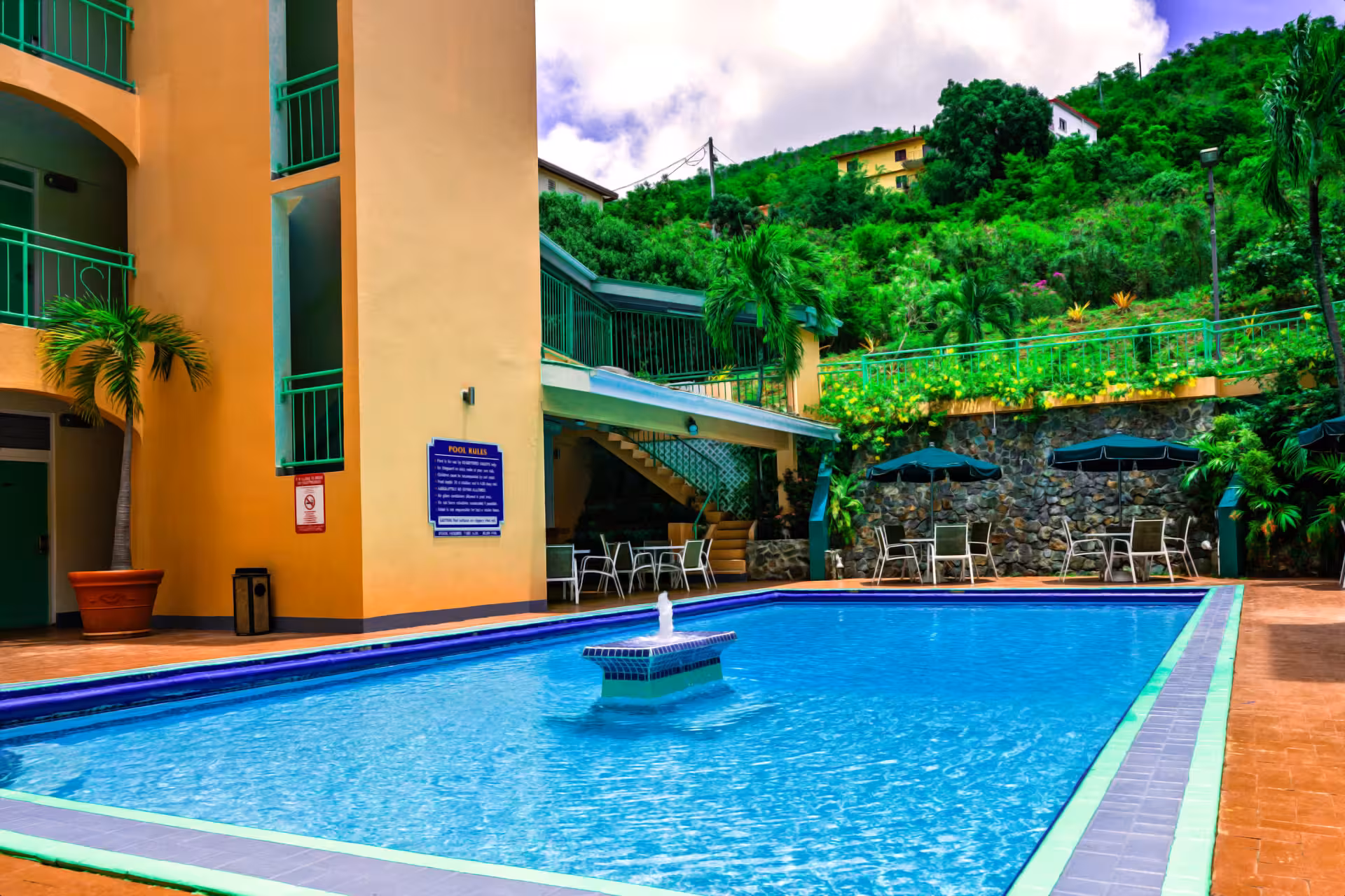 Outdoor swimming pool with a small fountain in the center, surrounded by patio tables with umbrellas and lush green hills in the background.