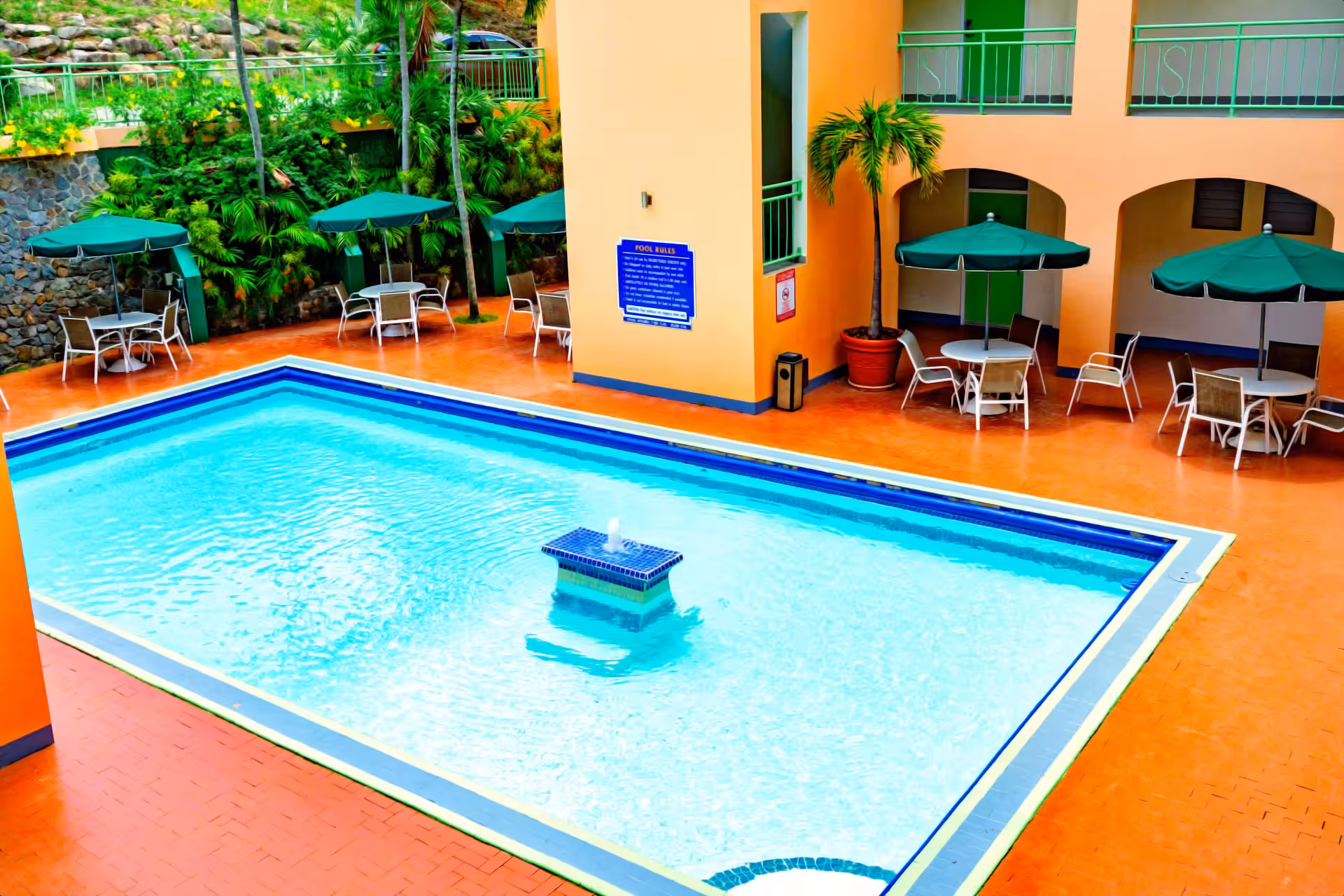 Outdoor rectangular swimming pool with clear water, surrounded by orange tiled patio, green umbrellas, white tables, chairs, and tropical plants.