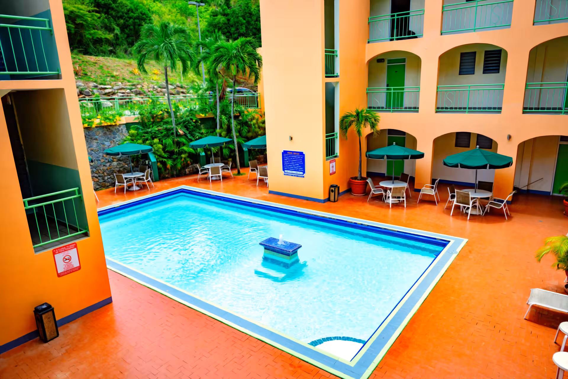 Rectangular outdoor swimming pool with a central water fountain, surrounded by orange-tiled flooring, green umbrellas, tables, chairs, and a peach-colored building with green railings.