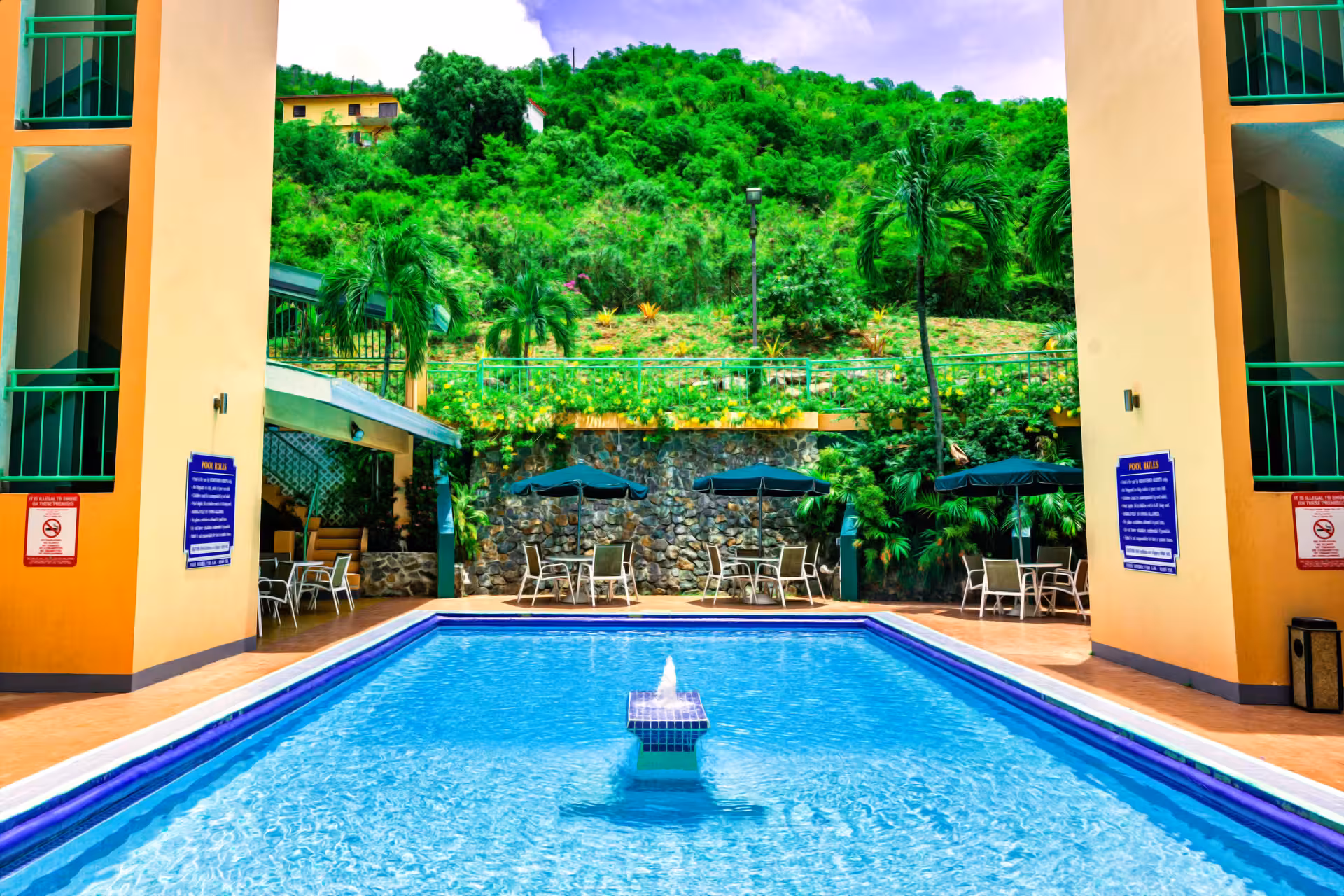 Outdoor swimming pool with fountain in the center, surrounded by chairs, tables with umbrellas, and tropical greenery.