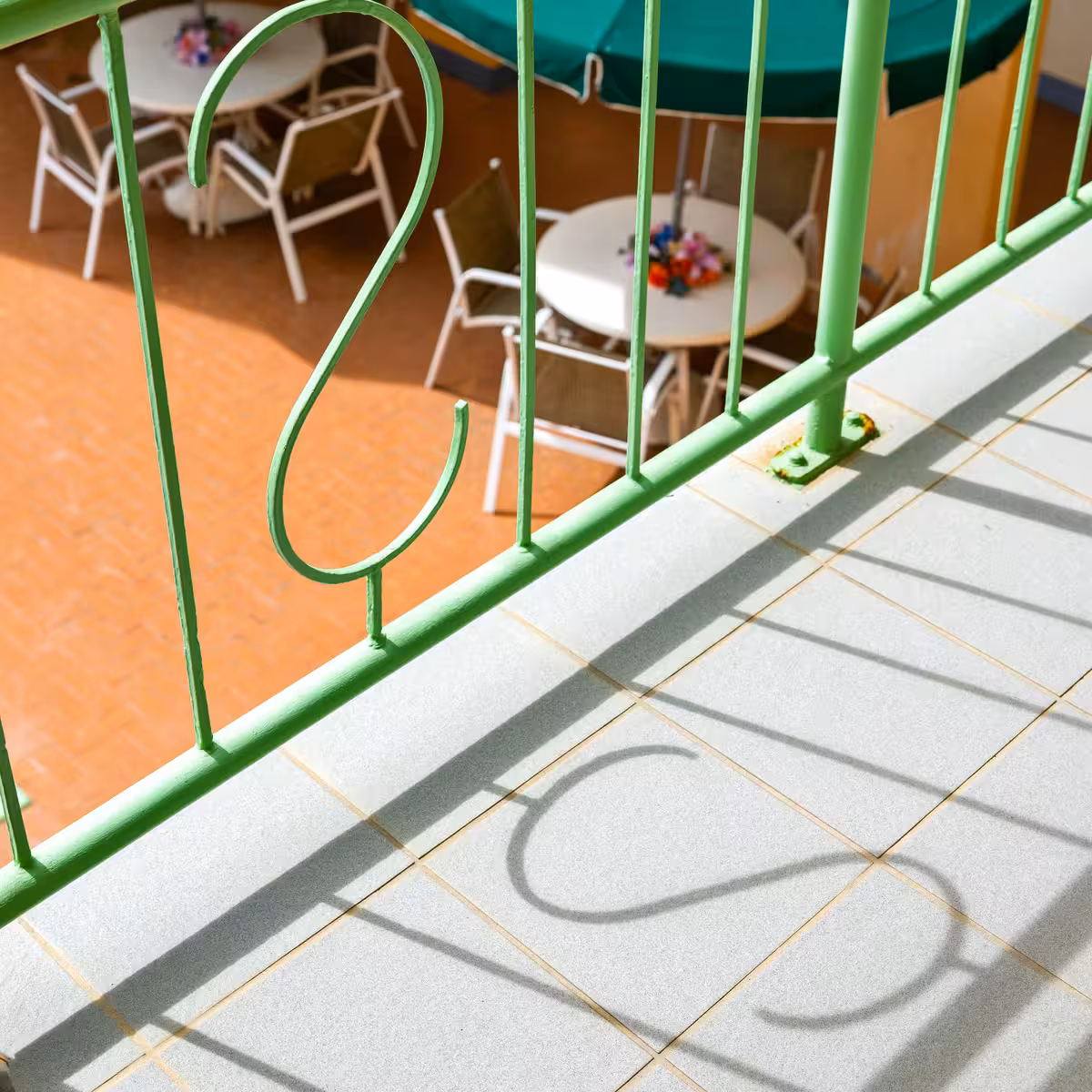 Green metal balcony railing casting shadow on tiled floor with outdoor tables and chairs below.