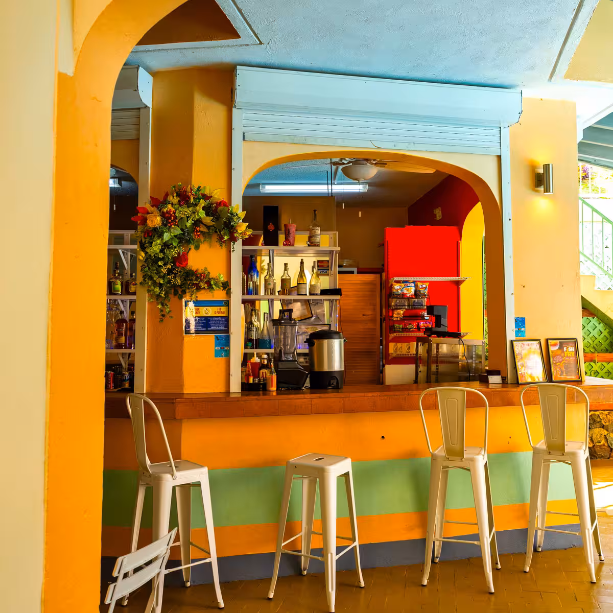 Interior of a colorful bar with orange and green walls, four white metal stools, and a counter with bottles and snacks behind an archway.