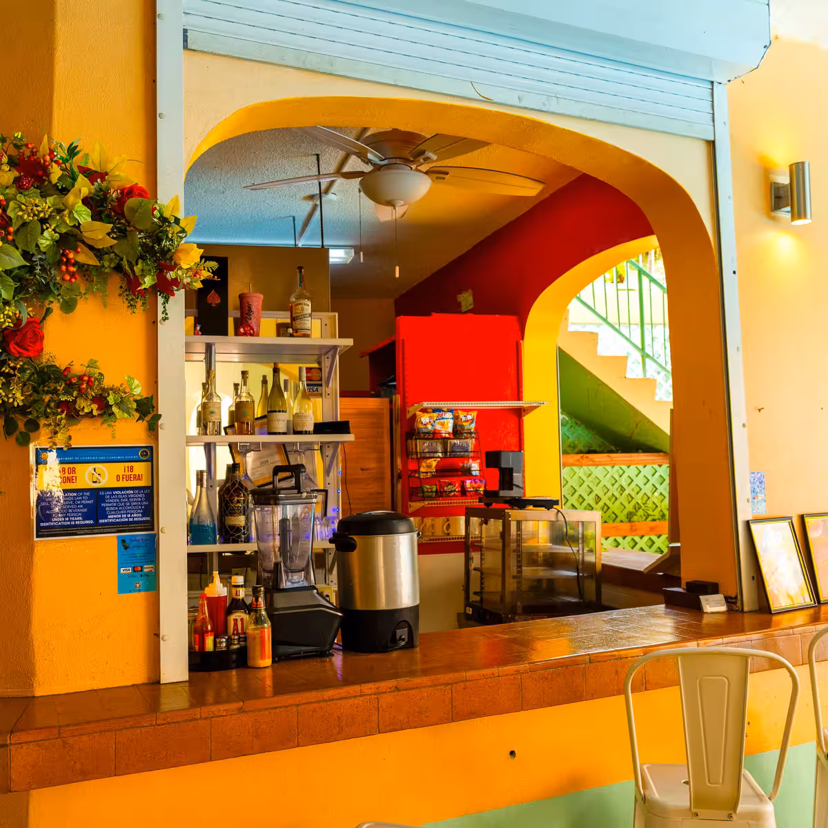 Colorful bar or café counter with bottles, blender, condiment bottles, and decorative flowers on the left wall.