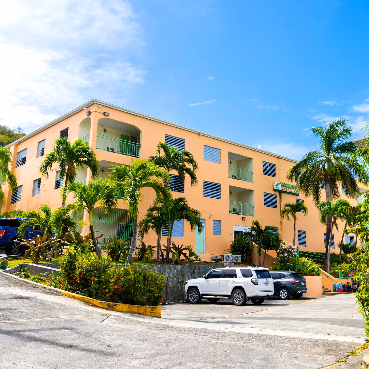 Three-story peach-colored hotel building with green railings, surrounded by palm trees, tropical plants, and parked cars under a bright blue sky.
