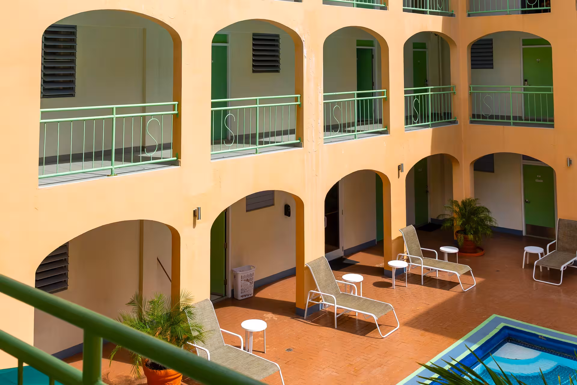 Hotel courtyard with peach walls, green doors, lounging chairs, potted plants, and a small blue swimming pool.
