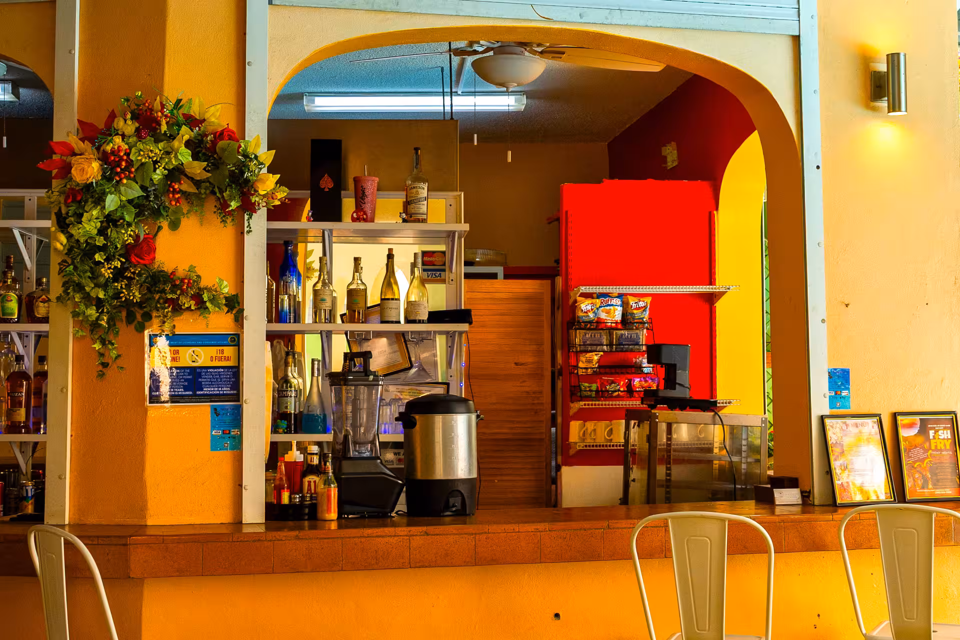 Colorful bar counter with bottles, a blender, beverage dispenser, snack rack, floral wreath, and three white metal chairs.
