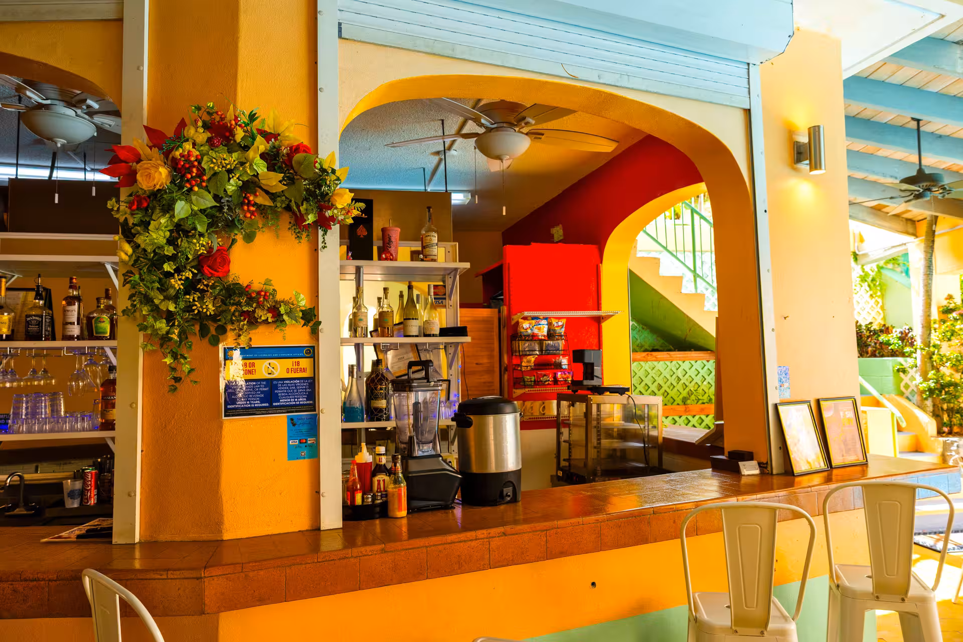 Colorful bar area with orange walls, floral wreath hanging on the left, bottles and blender on the counter, and white metal chairs.