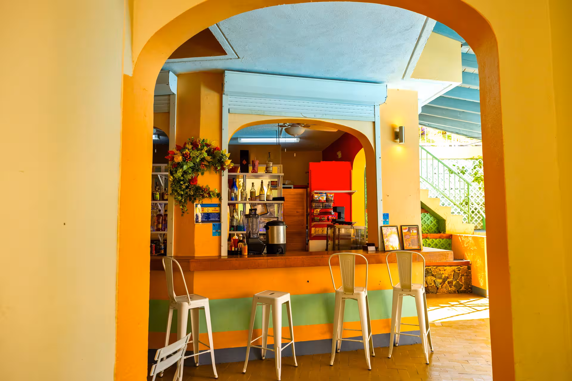 Vibrant yellow and green cafe counter with white metal stools, archways, and a decorated wall with bottles and snacks.