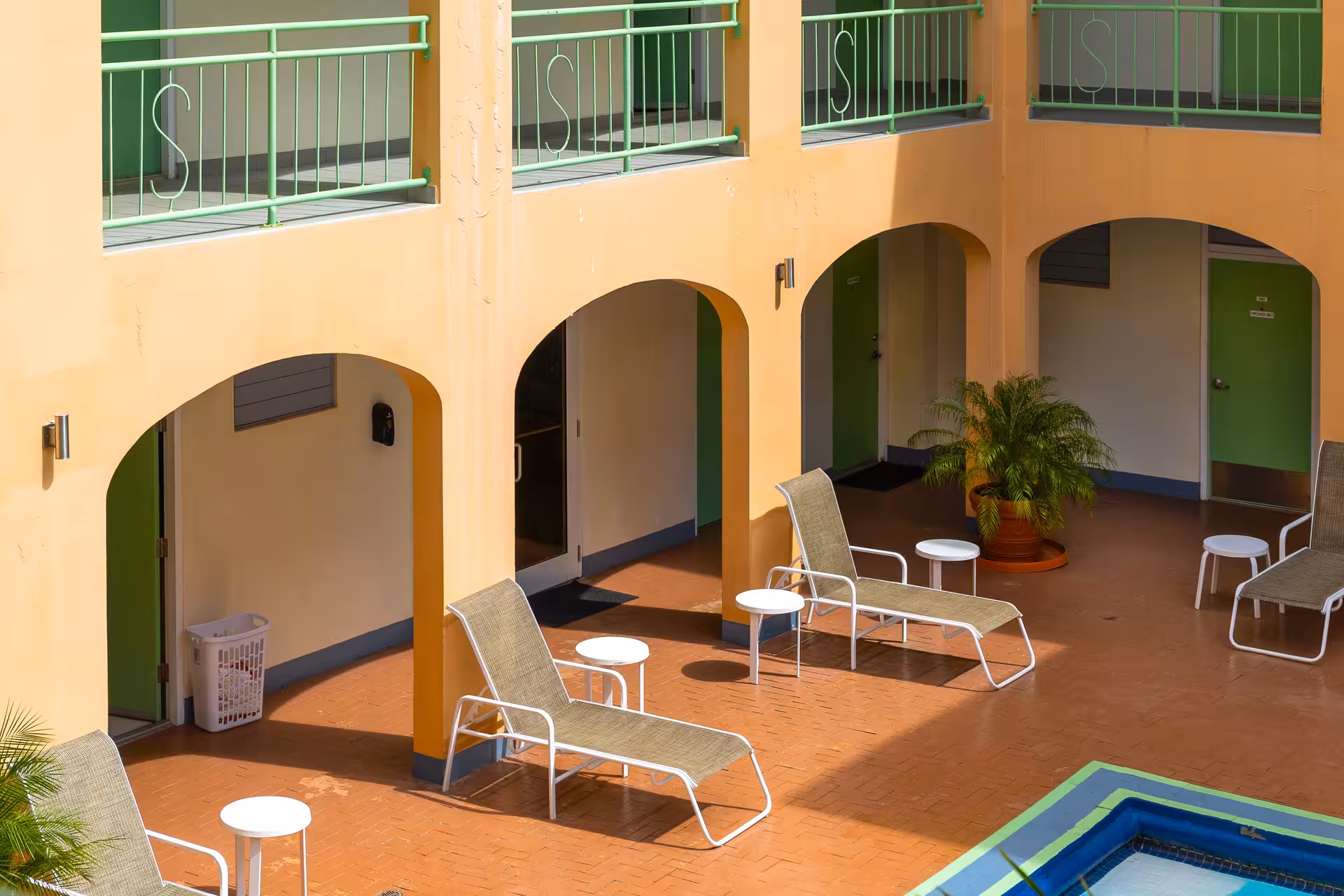 Poolside area with lounge chairs, small white tables, potted plants, and green doors in a peach-colored hotel courtyard.