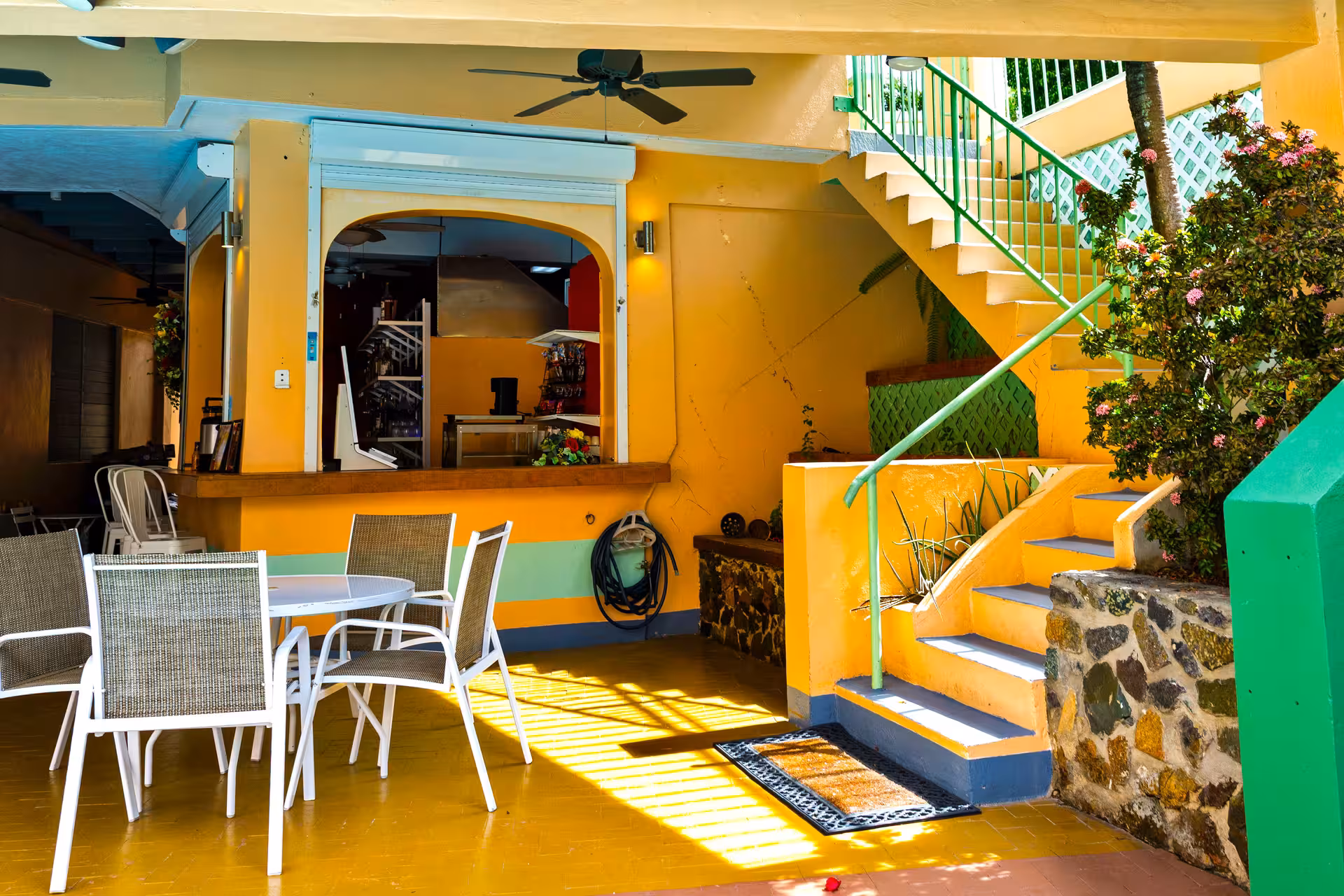 Outdoor seating area with white round table and four chairs next to yellow walls and stairs with green railing, plants, and flowers.