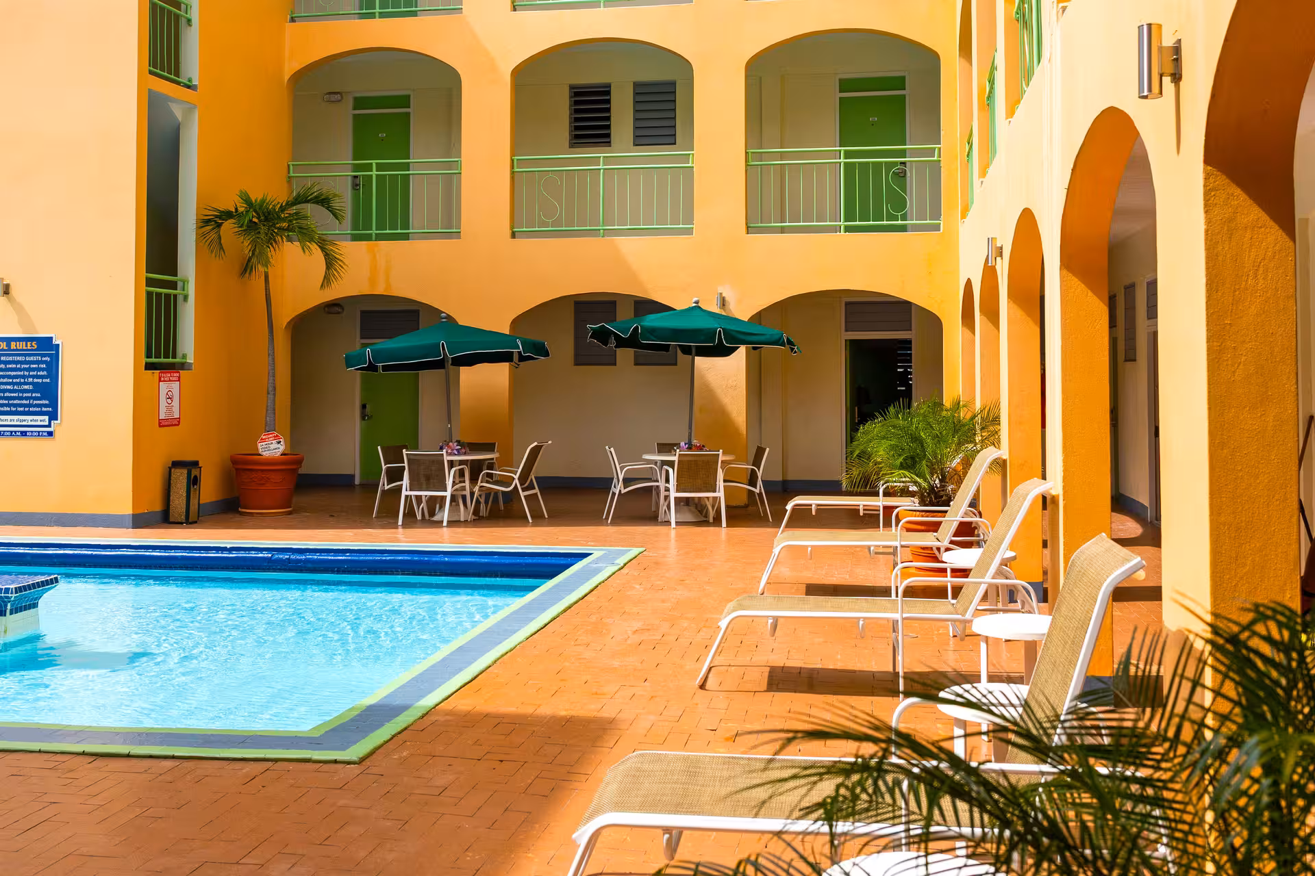 Outdoor hotel pool area with blue water, surrounded by orange tiled floor, lounge chairs, green umbrellas, and yellow building with green doors and arches.
