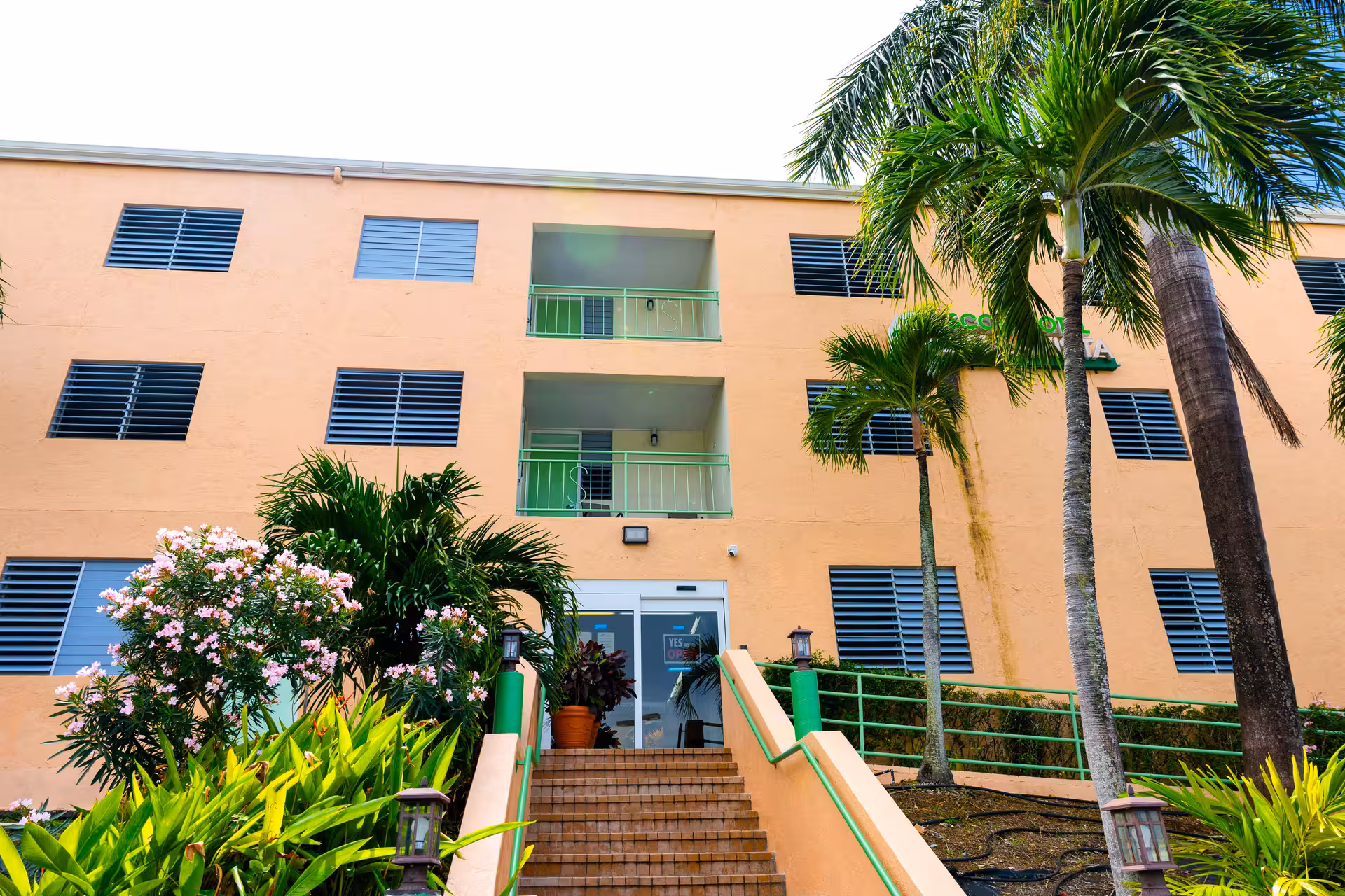 Orange hotel building with green railings, stairs leading to glass entrance doors, surrounded by palm trees and flowering plants.