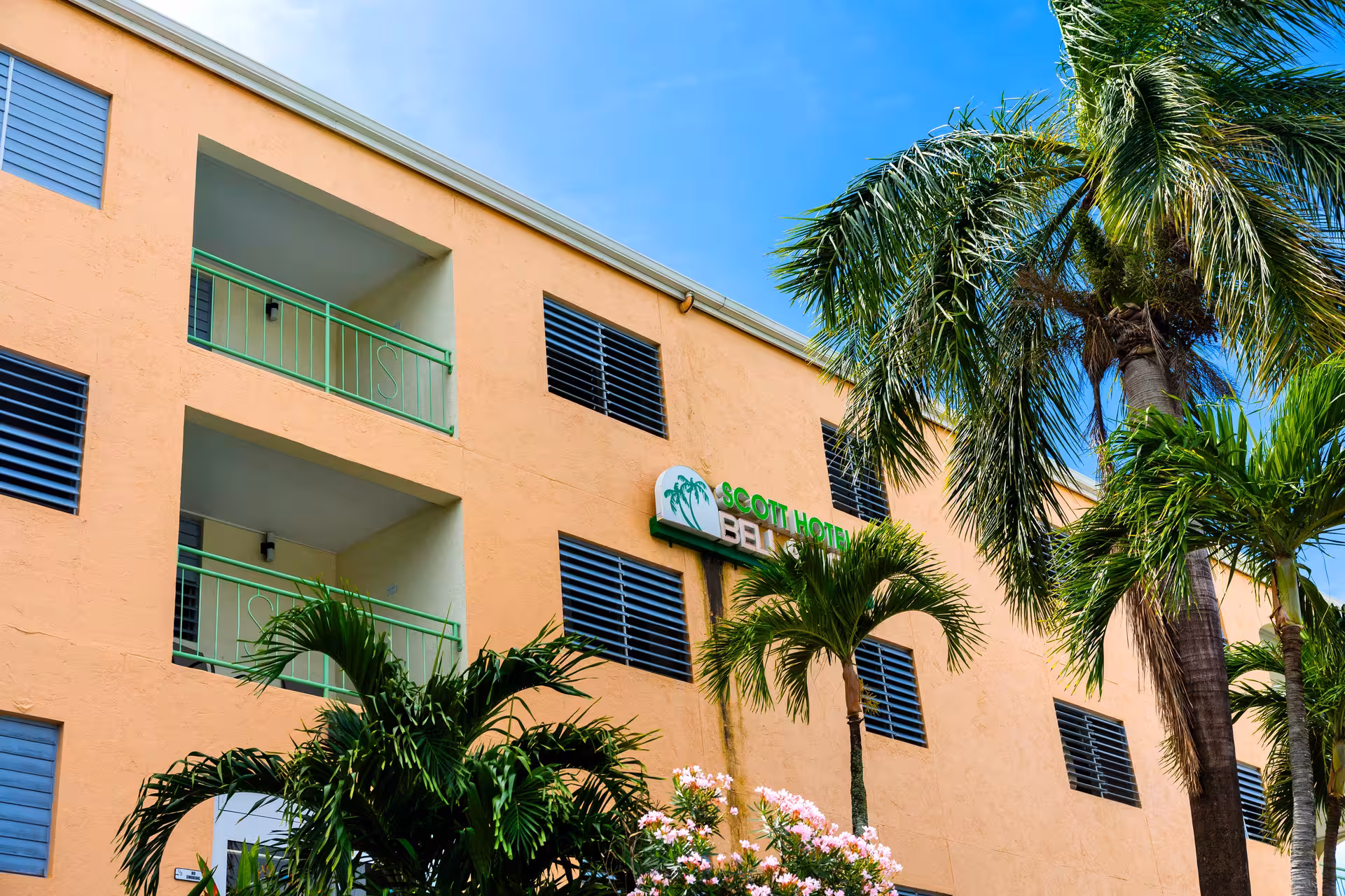 Exterior of a peach-colored Scott Hotel building with green railings on balconies and surrounded by palm trees under a clear blue sky.