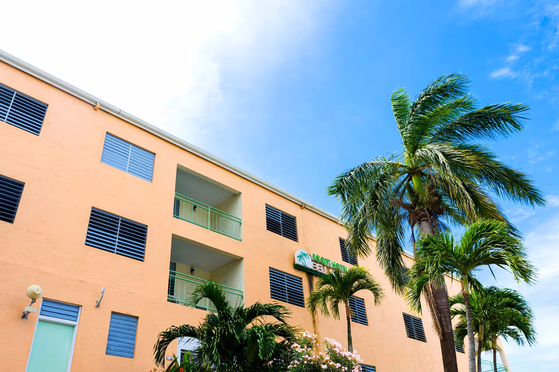 Peach-colored hotel building with green balconies, surrounded by palm trees against a blue sky.
