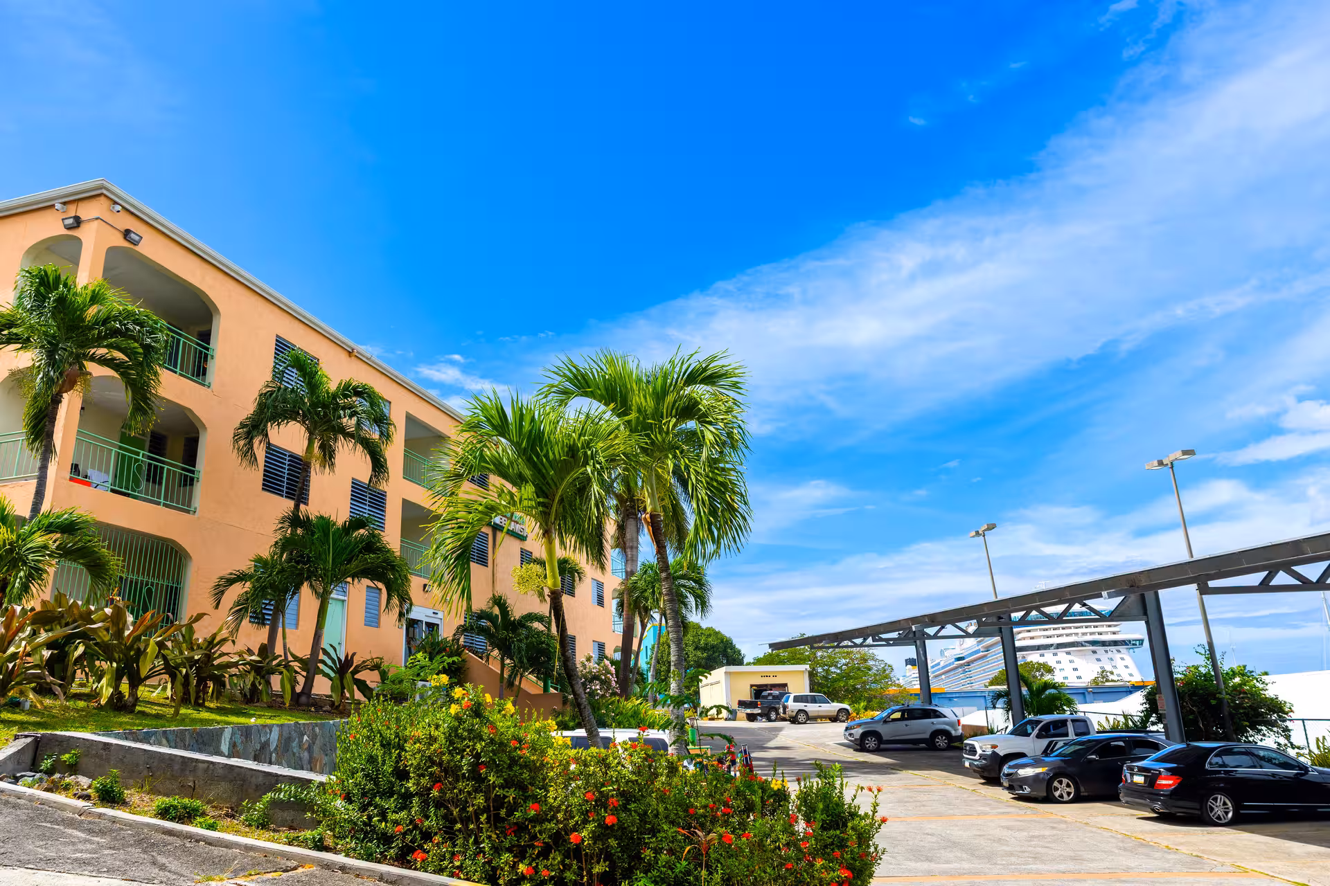 Exterior of a peach-colored building with green railings, surrounded by palm trees and bushes, with a parking lot and a cruise ship partially visible in the background under a blue sky.