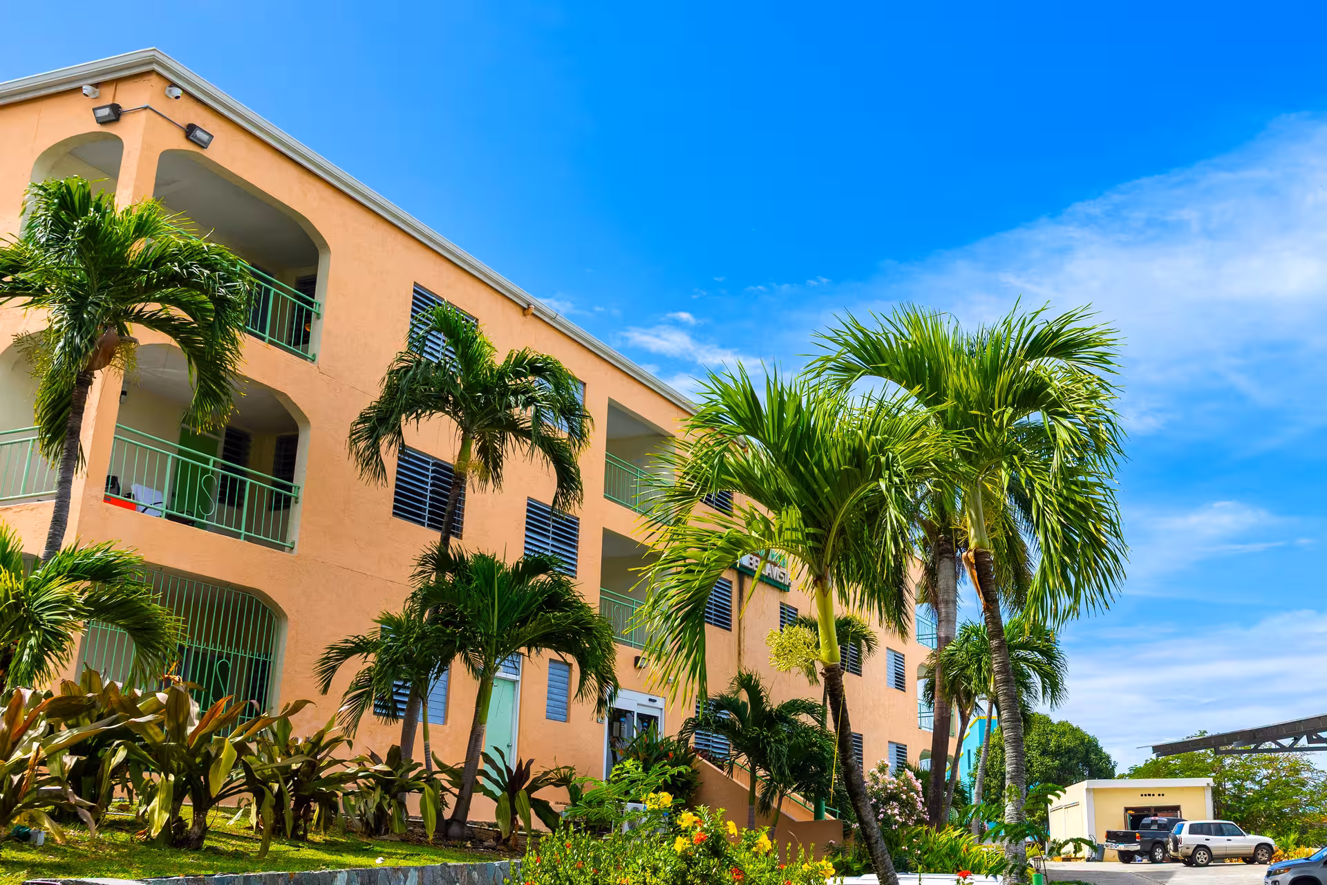 Peach-colored three-story hotel building with green balconies surrounded by palm trees and tropical plants under a bright blue sky.