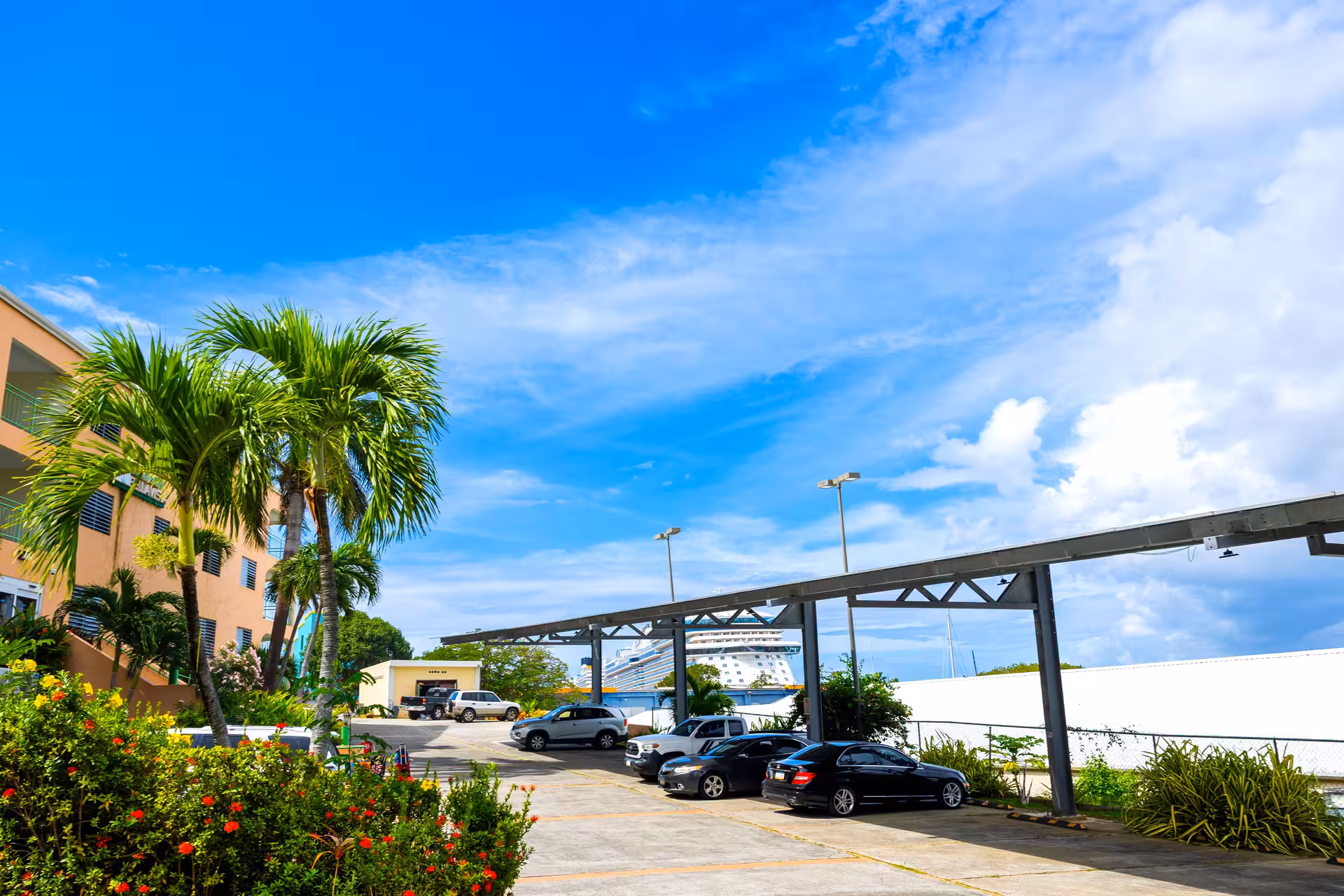 Parking lot with palm trees and colorful flowers beside a beige building under a bright blue sky with a cruise ship visible in the background.