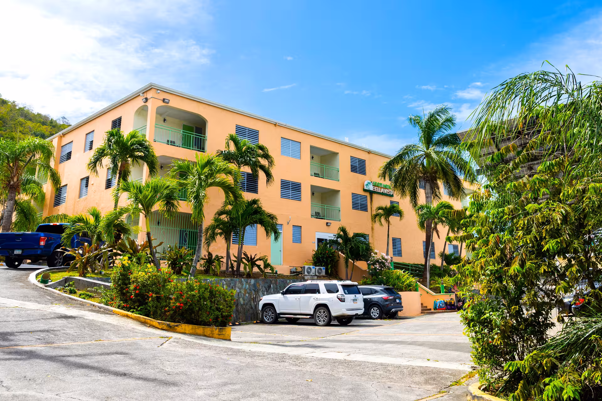 Three-story peach-colored hotel building with green railings surrounded by palm trees and parked cars under a blue sky.
