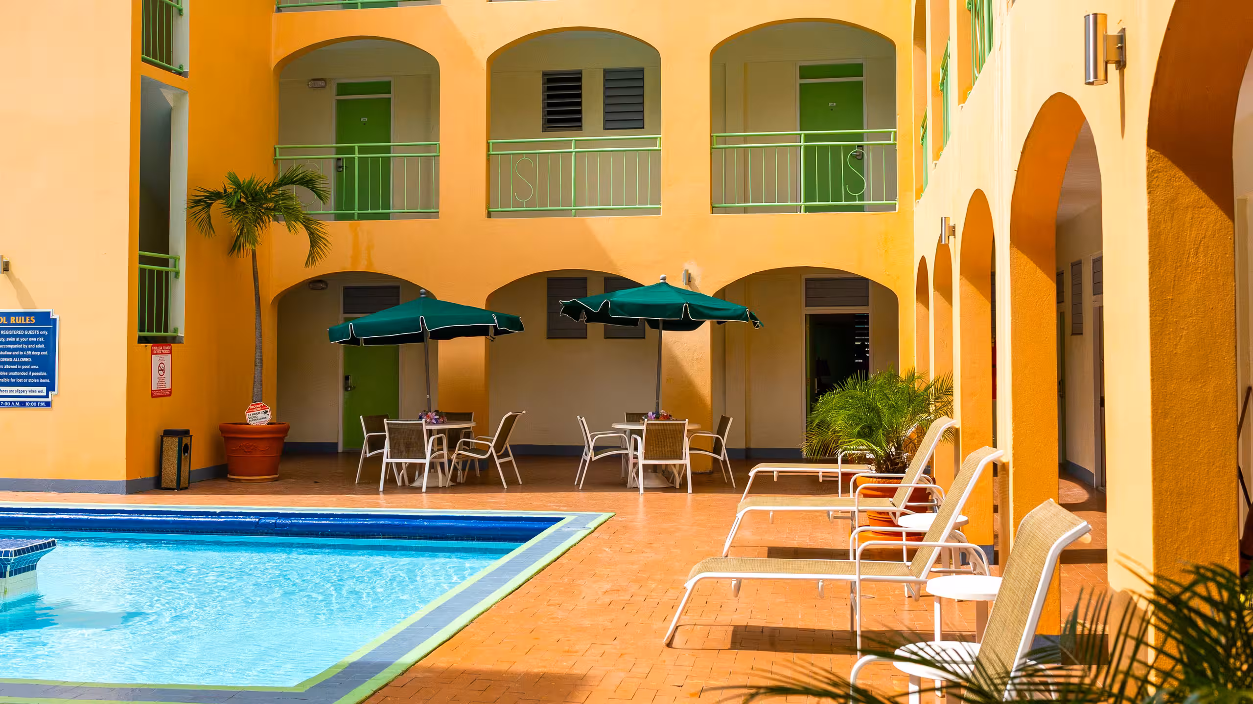 Hotel courtyard with a swimming pool, sun loungers, tables with green umbrellas, and orange walls with green doors and railings.