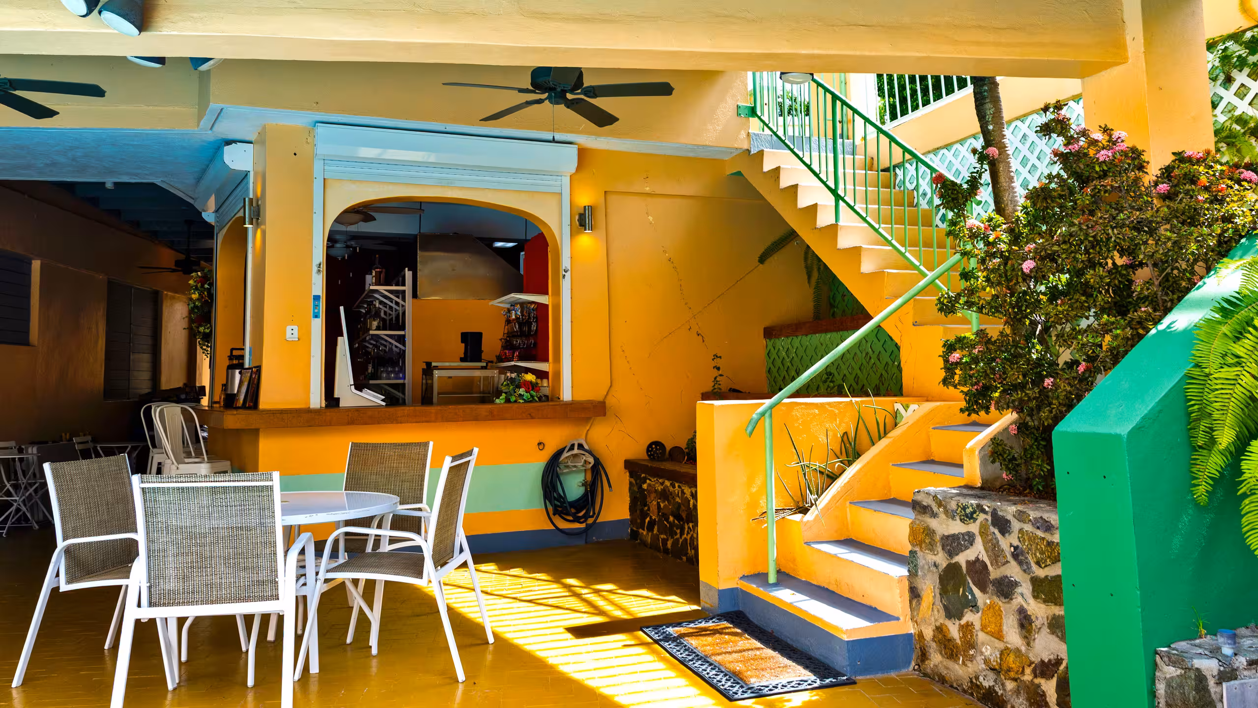 Outdoor seating area with white metal chairs and table next to yellow walls and stairs with green railings surrounded by plants.