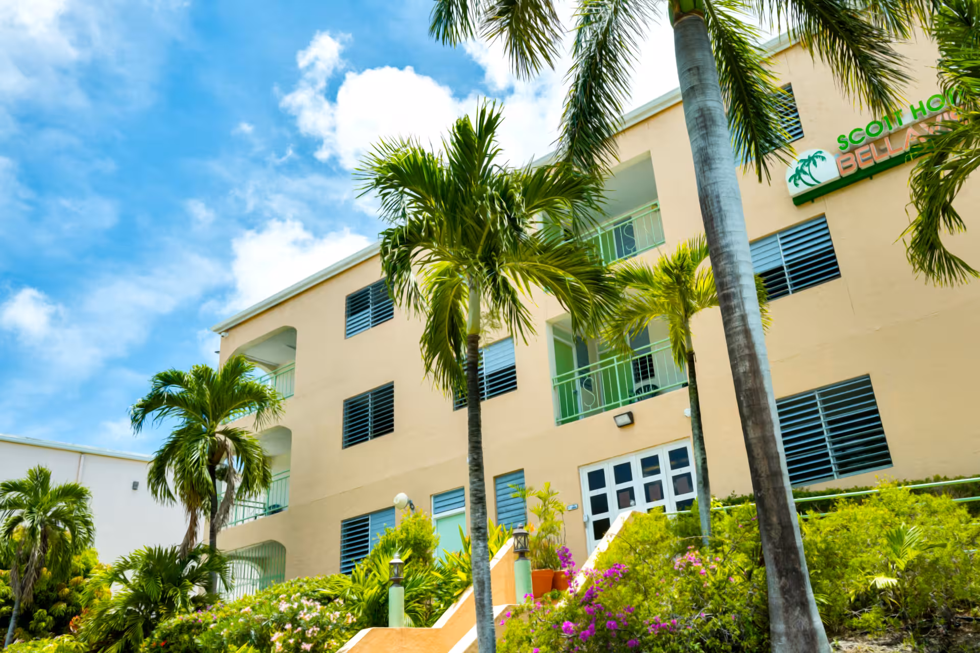 Exterior view of a beige multi-story building with green railings surrounded by palm trees and tropical plants under a partly cloudy blue sky.