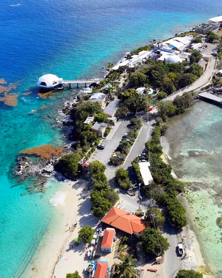 Aerial view of a coastal park with turquoise waters, a white dome structure on a pier, sandy beach, buildings with red roofs, parking areas, and lush greenery.