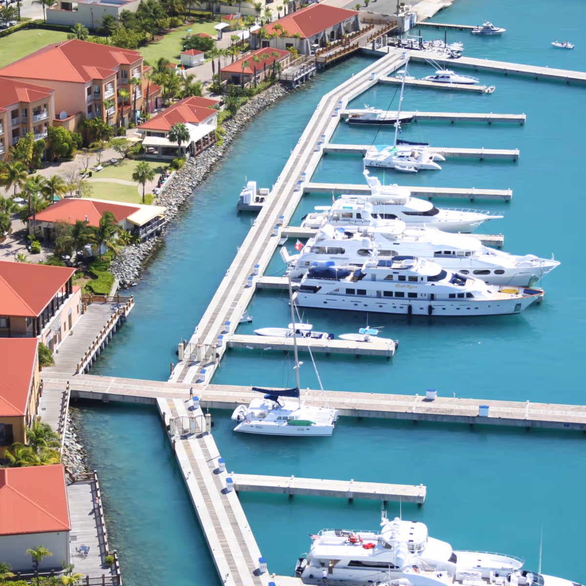 Aerial view of a marina with multiple white yachts docked alongside piers, red-roofed buildings, and turquoise water.