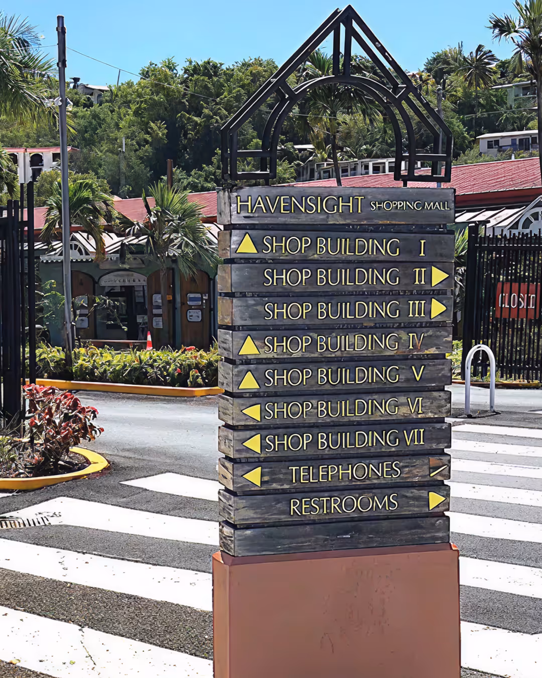 Wooden signpost at Havensight Shopping Mall showing directions to shop buildings I to VII, telephones, and restrooms with arrows.