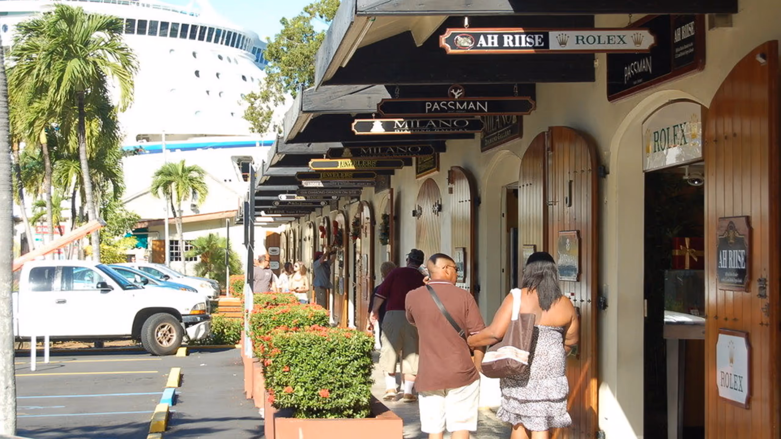 People walking along a shopping arcade with wooden doors and hanging store signs, palm trees, parked cars, and a large cruise ship in the background.