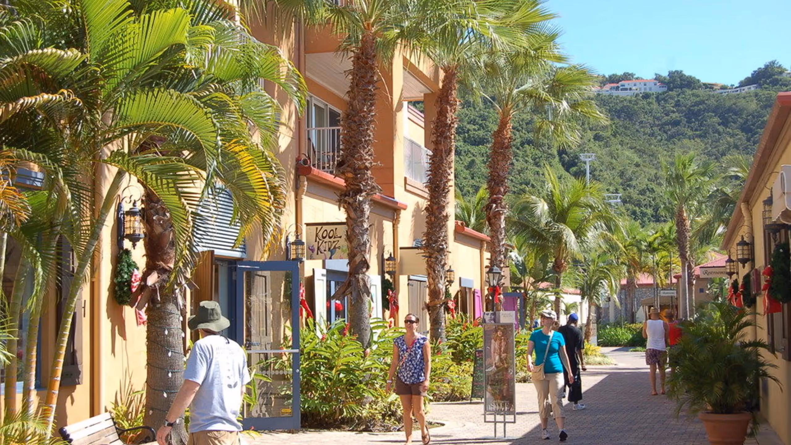 People walking along a sunny, palm-lined outdoor shopping area with yellow buildings and green hills in the background.