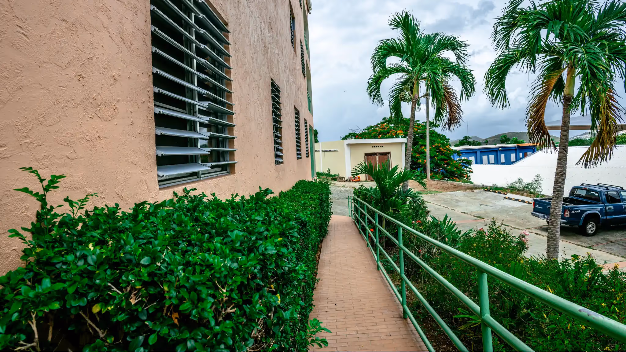 Accessibility ramp with green metal railing alongside a beige building with louvered windows, surrounded by green bushes and palm trees.