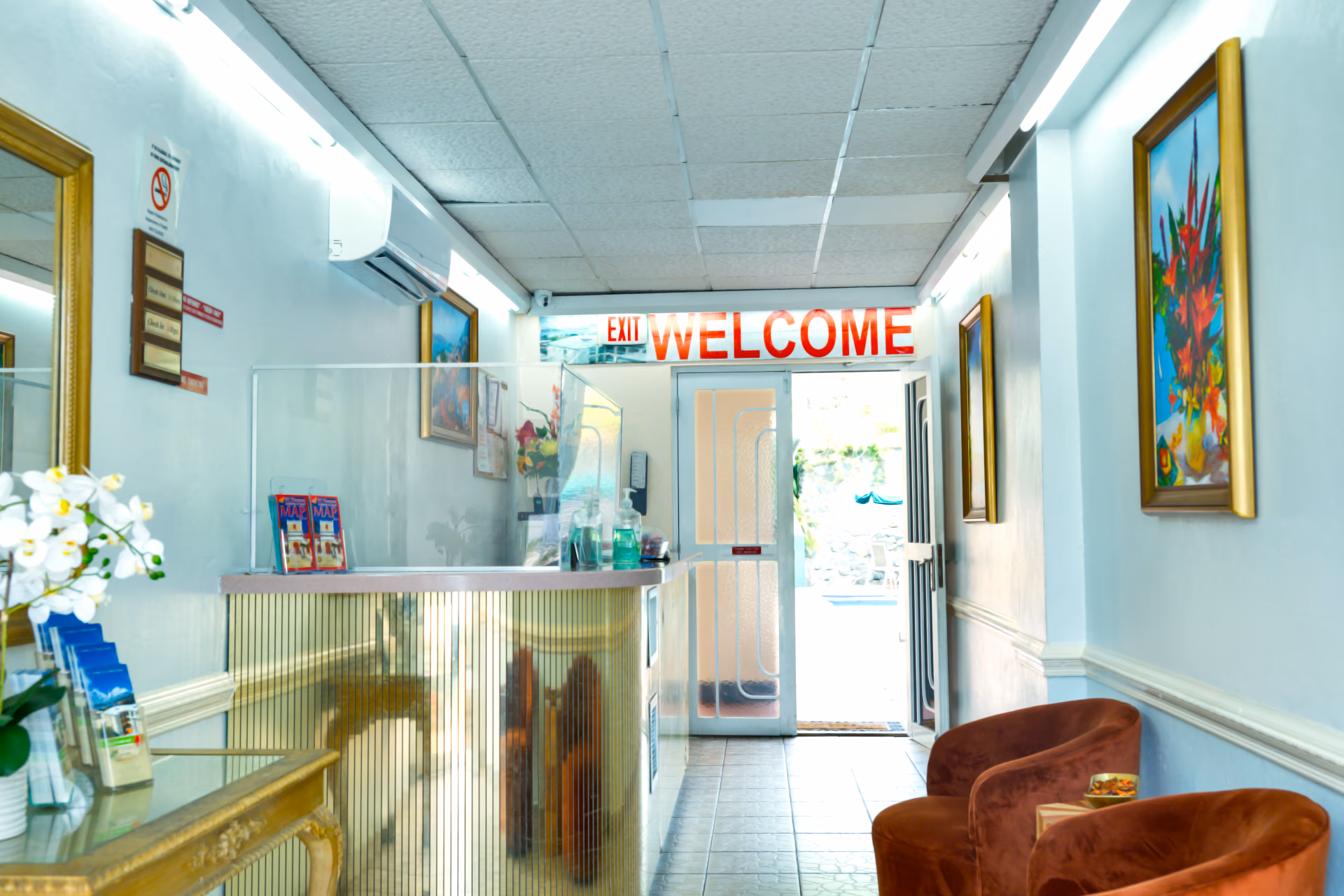 Bright reception area with a welcome sign above a glass door, two brown chairs, framed paintings, and a front desk with brochures and hand sanitizer.