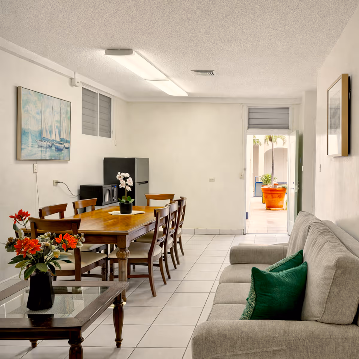 Bright dining and living area with wooden table and chairs, gray sofa with green pillows, and decorative flowers.