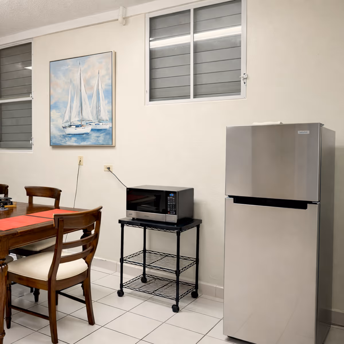 Dining area with wooden chairs, a table with a red runner, a microwave on a black metal rack, and a stainless steel refrigerator against a white wall with a painting of sailboats and two windows.