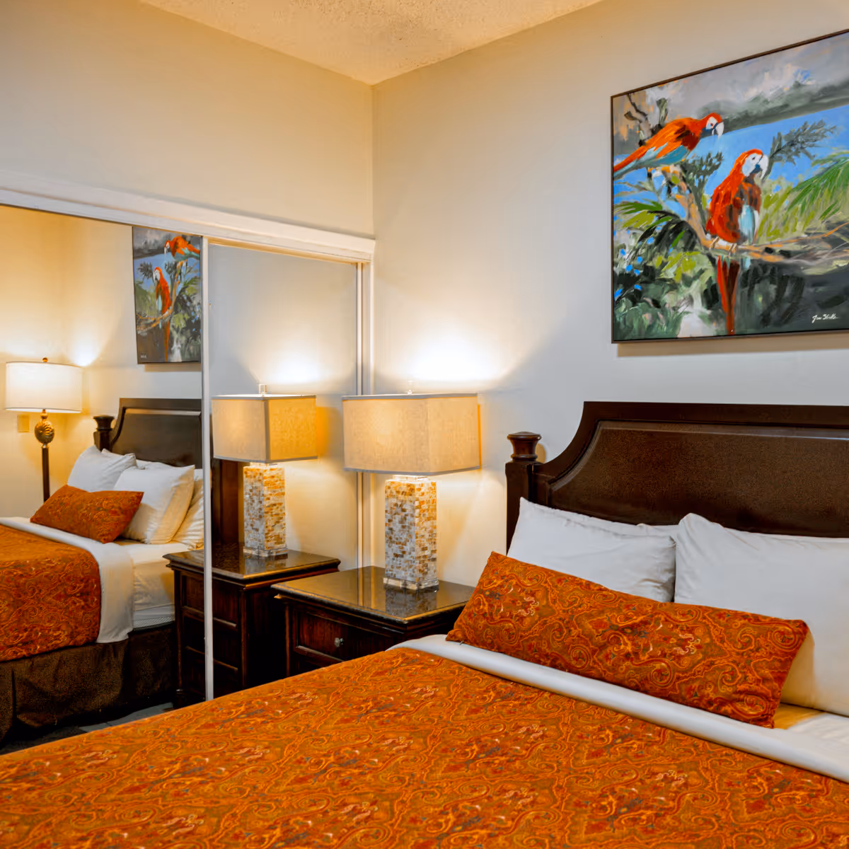 Cozy hotel bedroom with a dark wooden headboard, white pillows, orange patterned bedspread and pillow, bedside table with textured lamp, and painting of two red parrots on the wall.