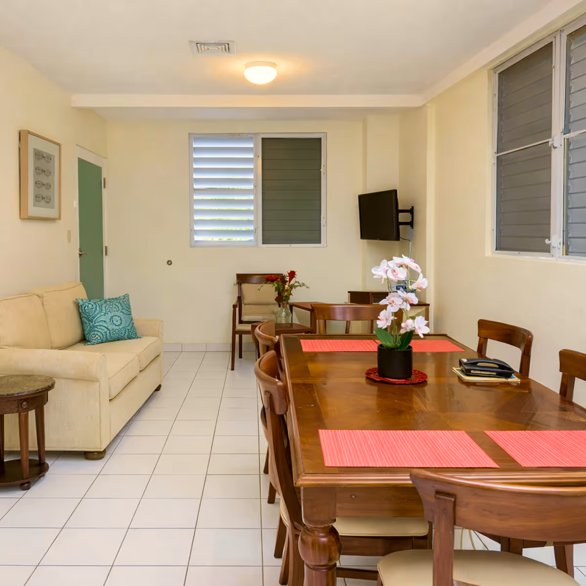 Bright dining and living area with a wooden dining table set with red placemats and a floral centerpiece, adjacent beige sofa with a turquoise pillow.
