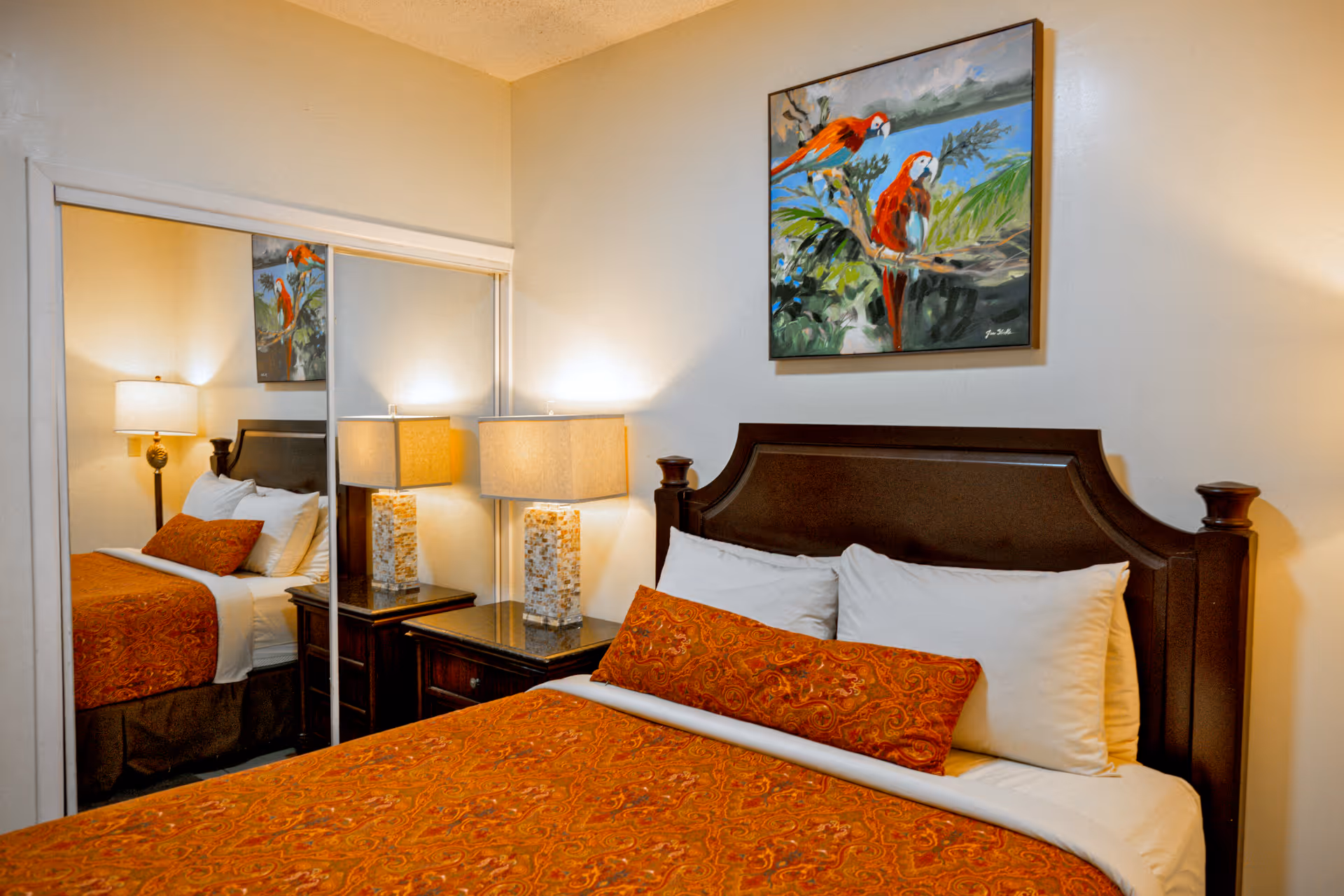 Bedroom with dark wooden headboard, orange patterned bedspread and pillow, two white pillows, a bedside table with a mosaic lamp, and a painting of two red parrots on a branch above the bed.