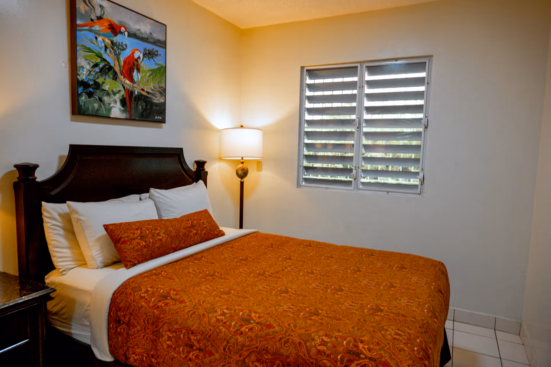 Bed with ornate wooden headboard, orange patterned bedspread, white pillows, standing lamp, and window with horizontal blinds in a bedroom.