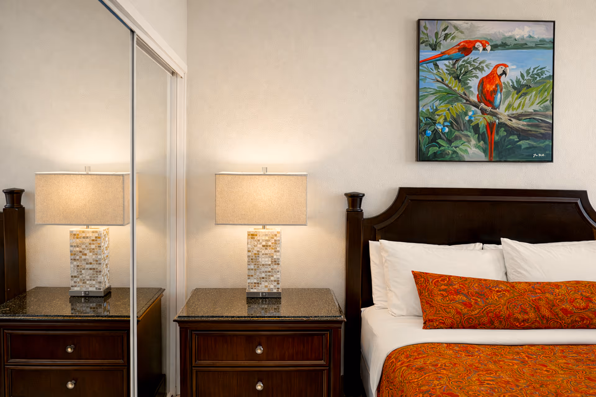 Bedside table with mosaic lamp next to a dark wood bed with white pillows and orange patterned bedding, under a painting of red parrots.