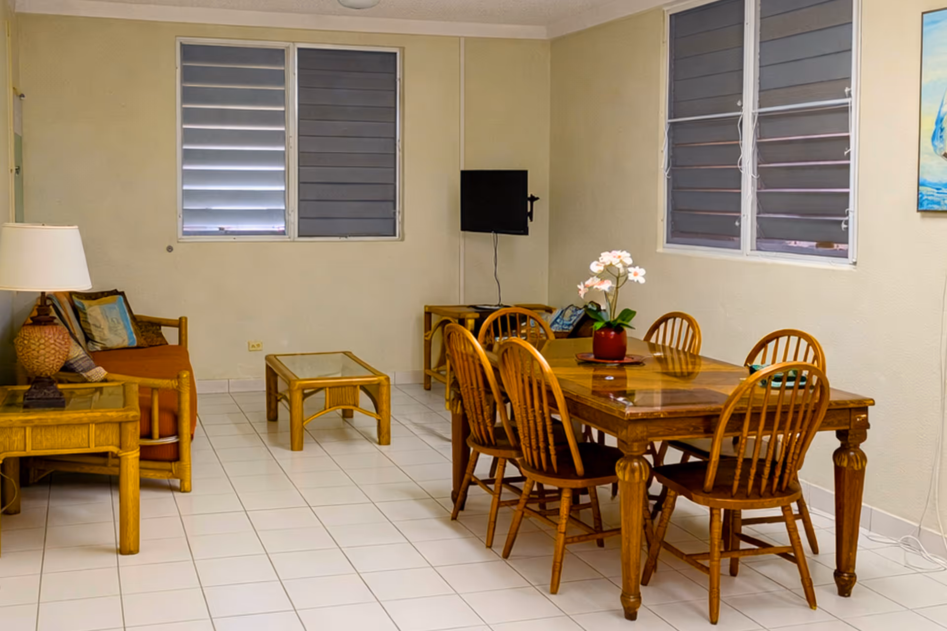 Living and dining area with wooden dining table and six chairs, a small sofa, side tables, and a wall-mounted TV under shuttered windows.