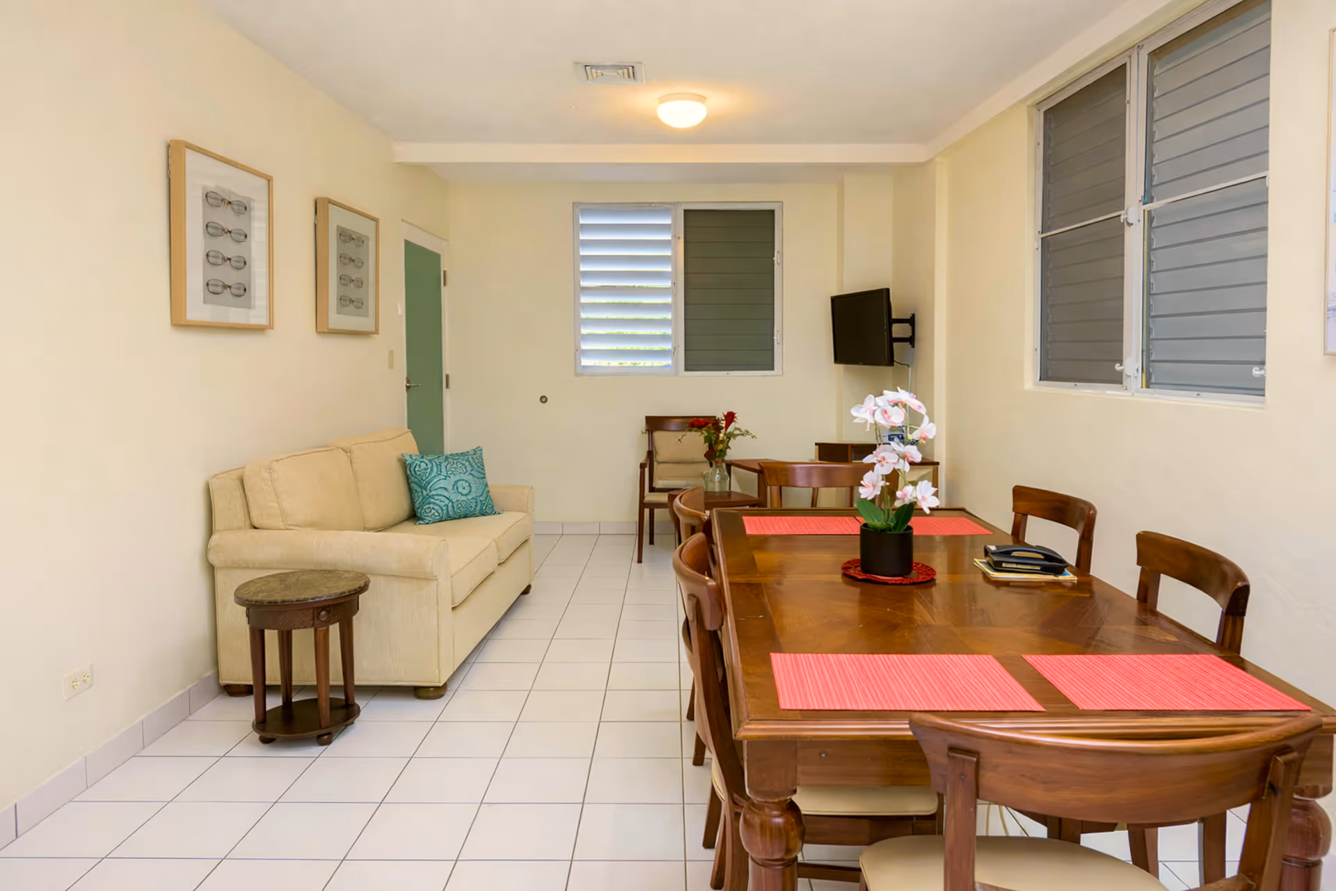 Bright dining room with wooden table and chairs, pink placemats, orchid centerpiece, beige sofa, side table, and wall-mounted TV.
