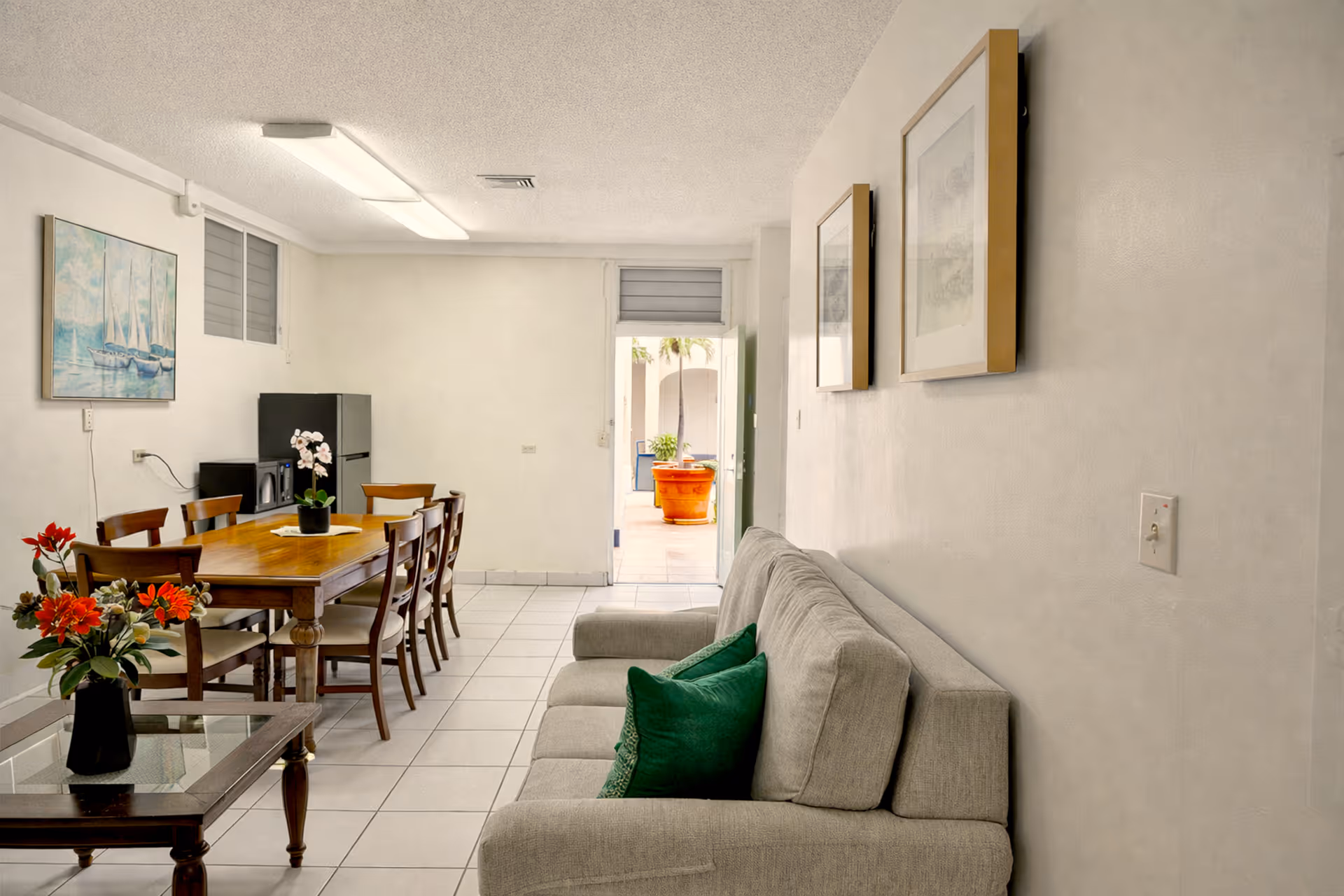 Bright living and dining area with a gray sofa with green pillows, wooden dining table with six chairs, and decorative flowers on tables.