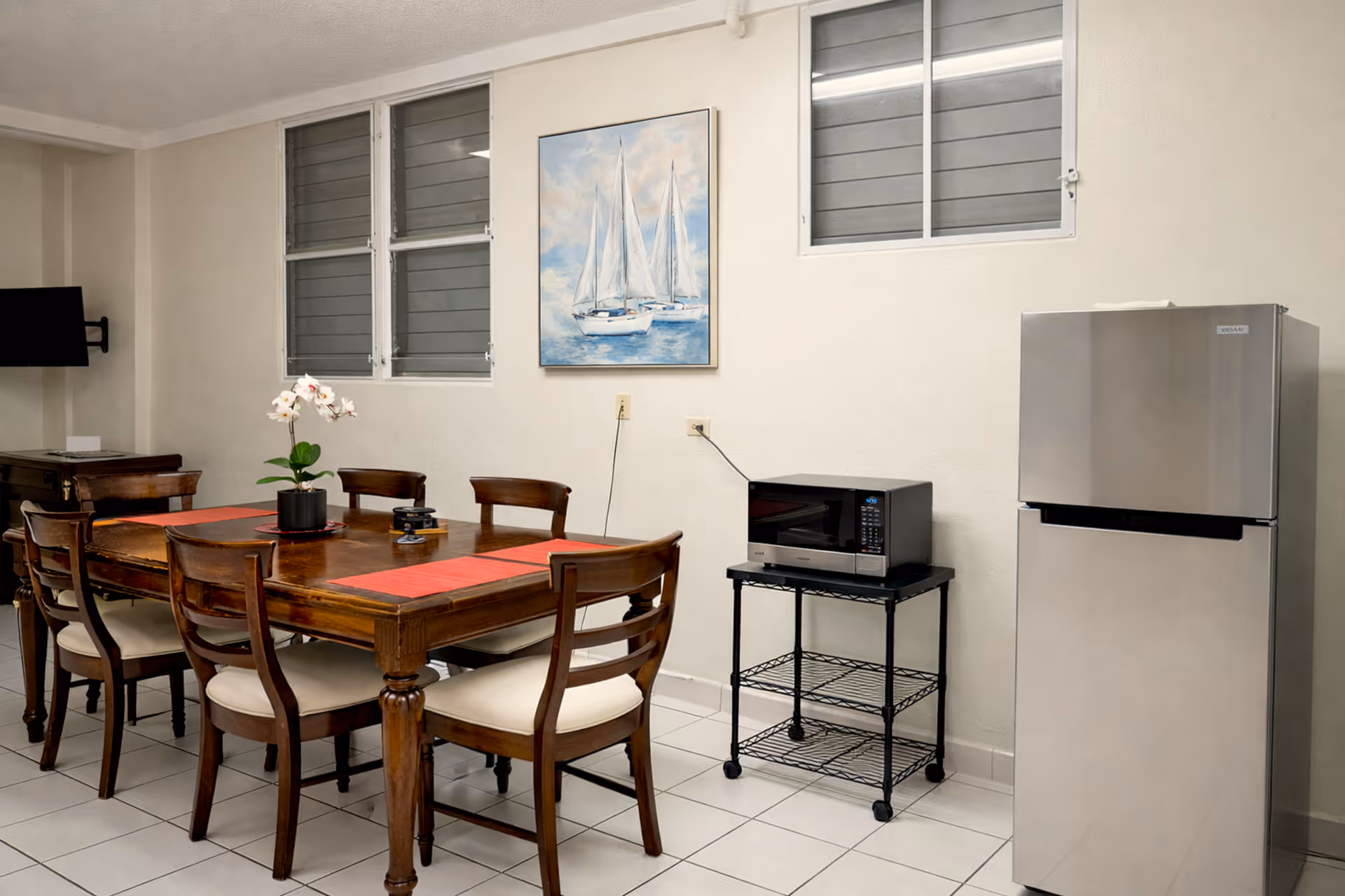 Dining area with a wooden table set for six, an orchid centerpiece, a microwave on a black metal cart, and a stainless steel refrigerator.